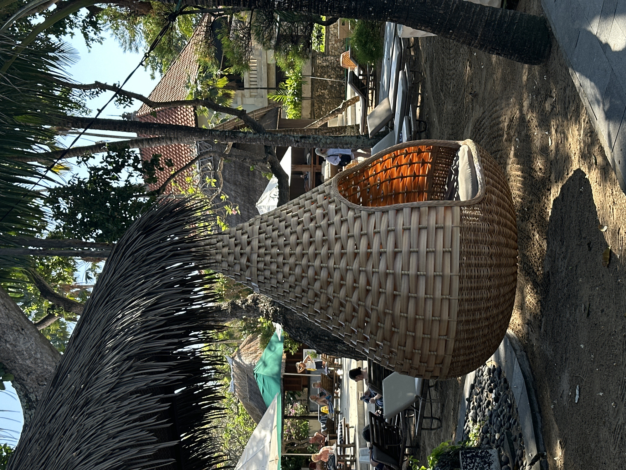 The image depicts an outdoor scene at what appears to be a resort or beachside establishment in Denpasar, Indonesia. In the foreground, a large, teardrop-shaped woven hanging chair with an orange interior and a gray cushion is suspended, casting a shadow on the sandy ground. To the left, a thatched roof structure extends into the frame. Further into the background, several lounge chairs are arranged on the sand, some occupied by people. Umbrellas, one white and one green, provide shade over seating areas. Behind these, a multi-story building with a terracotta tiled roof is visible, surrounded by lush green foliage and trees, including palm trees. The sky is clear and blue. The overall environment suggests a tropical vacation setting.