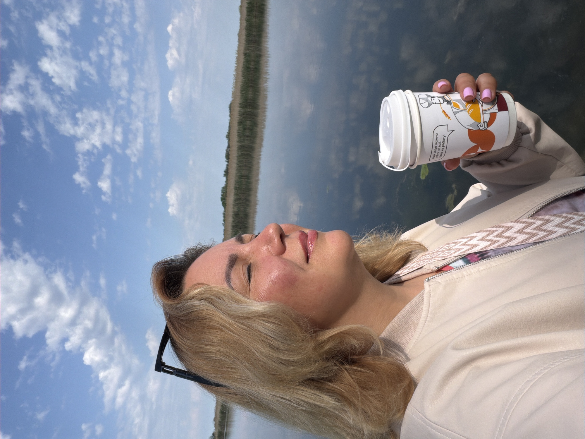 The image shows a woman holding a disposable coffee cup outdoors. She is blonde and wearing a beige jacket. The background is a lake with reflections of the sky and trees. The sky is blue with scattered clouds. The text on the coffee cup is in Russian and includes a cartoon image. The location is likely in Leningradskaya, Russia, based on the presence of the Russian text.