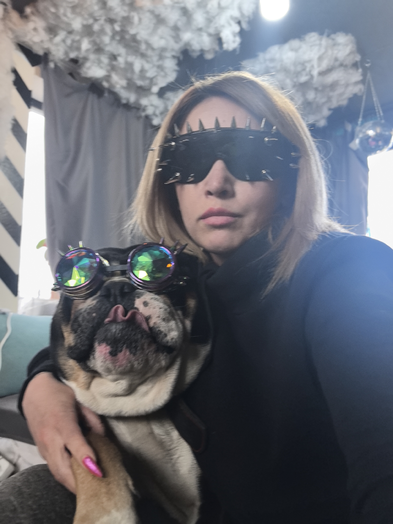 A woman and an English Bulldog are the focus of this selfie-style photograph taken indoors. The woman is wearing black spiked sunglasses and a black turtleneck sweater. She is holding the dog, who is also wearing sunglasses, with silver rims and multi-colored lenses, and sticking its tongue out. The background consists of decorative cotton cloud-like fixtures hanging from the ceiling, grey curtains, a black and white striped wall, and a glimpse of a disco ball. The location appears to be in Noviy, Russia.