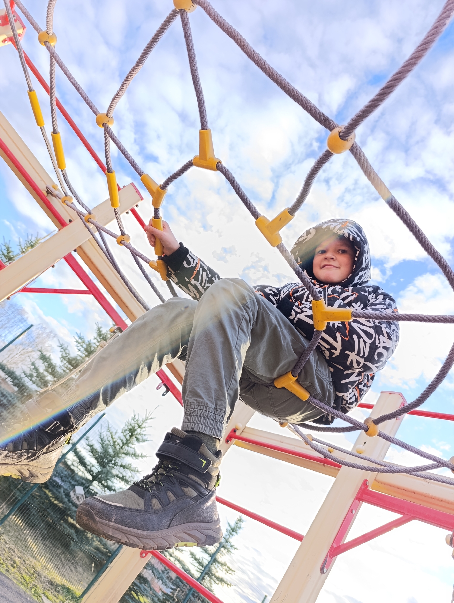 A low-angle shot captures a child on a playground climbing structure. The child, wearing a hooded jacket with a black and white graffiti-style pattern, green pants, and hiking boots, is positioned within a rope climbing net with yellow connectors. The net is anchored to a structure with red and off-white supports. The background shows a partly cloudy sky and hints of vegetation and fencing, suggesting an outdoor setting possibly in Фоки, Russia. The angle of the shot emphasizes the height and perspective of the child on the climbing structure.