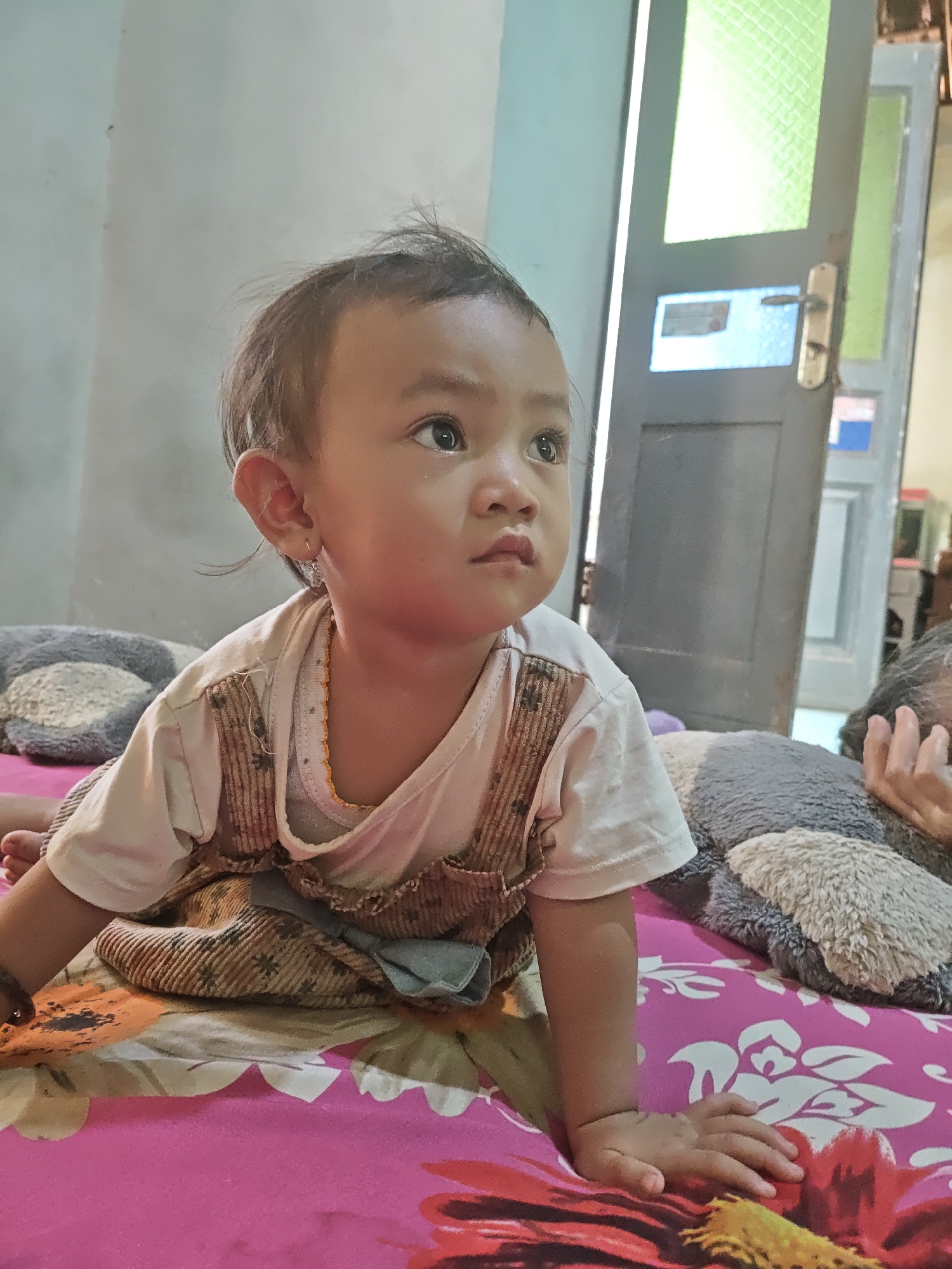 A close-up, eye-level shot shows a young child on a pink bed with a floral pattern in Ngasem, Indonesia. The child, with short, dark hair and brown eyes, is wearing a white t-shirt and a brown overall. The child is on their hands and knees, looking to the right with a thoughtful expression. The background includes a light-colored wall and a partially visible gray door with green glass panels, possibly a room in a house. The bed is covered with a pink floral sheet and a gray plush toy is visible to the right of the child. An adult's hand with grey hair is also visible on the right side of the image, resting on the plush toy.