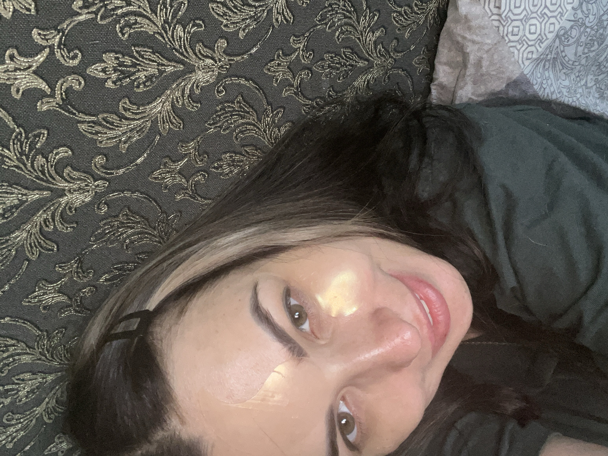 A young adult female is depicted in a close-up, diagonal shot, appearing to be lying down and looking towards the camera with a gentle smile. Her face is in the lower right portion of the frame, angled upwards. She has long, dark hair with lighter highlights or strands visible near her face, held back by a black bobby pin near her temple. Her eyebrows are dark and well-defined, and her eyes are brown. Clear or translucent facial patches, possibly hydrocolloid patches for skincare, are visible on her forehead and left cheek, reflecting some light. Her lips are a natural pink, slightly parted to reveal a hint of her teeth.
The setting appears to be indoors, possibly a living room or bedroom. To her left and behind her head is a dark fabric with an intricate, raised metallic gold or bronze floral and scroll pattern, suggestive of upholstery on a couch or headboard. To her right and below her, a plain dark fabric, likely her clothing (a dark shirt or blanket), is visible. In the upper right corner of the image, another piece of fabric, light gray with geometric and subtle paisley patterns, suggests a pillow or another blanket. The overall mood conveyed is relaxed and calm. There is no visible text in the image.