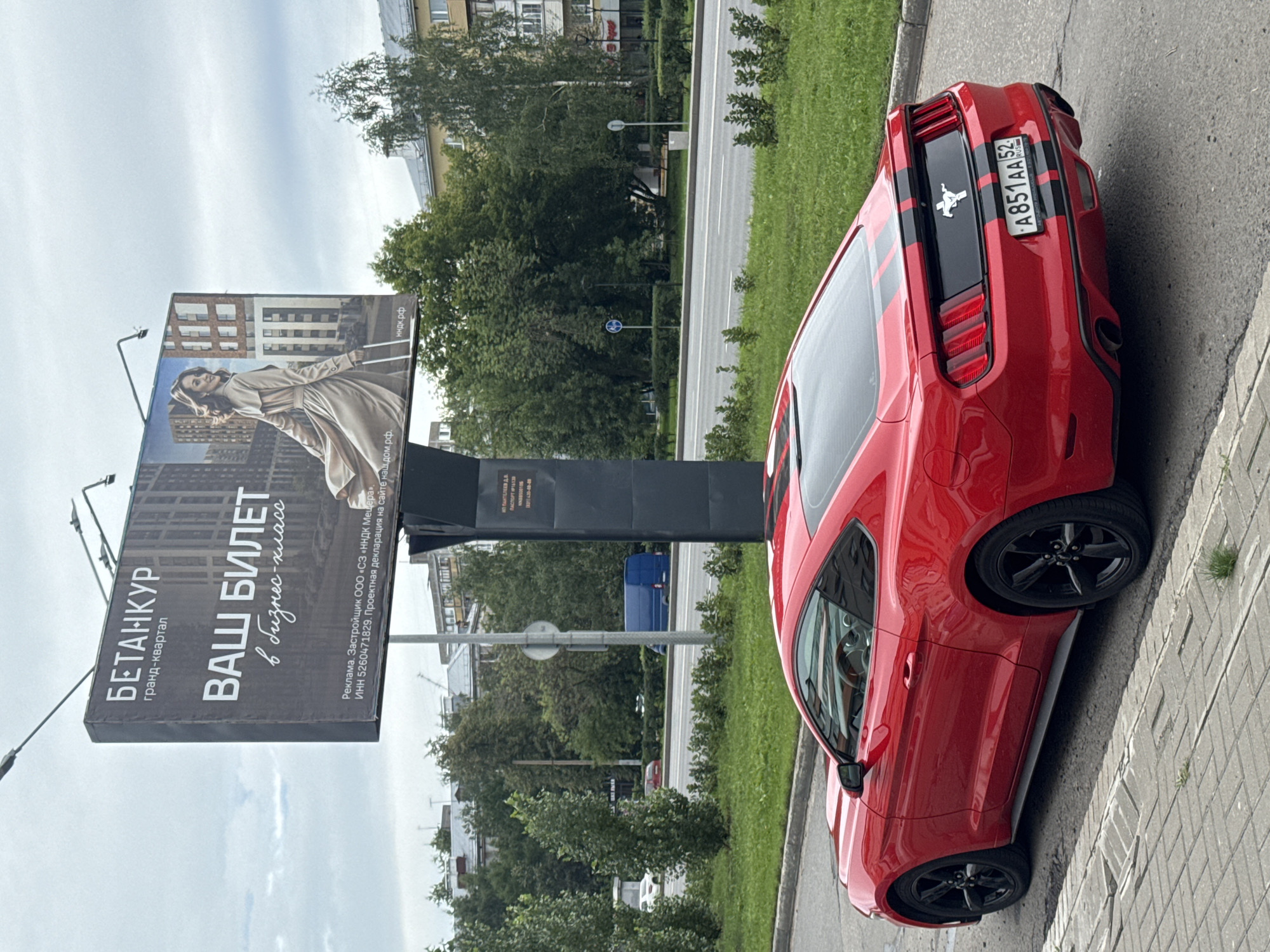 A red Ford Mustang is parked on the side of a street in Nizhny Novgorod, Russia. The car is a coupe with black racing stripes on the hood and trunk. The license plate reads "A851AA 52 RUS". The car is parked on a paved sidewalk, with a grassy area and a road beyond. In the background, there is a large billboard advertising a residential complex called "Betankur Grand-kvartal". The billboard features an image of a woman in a trench coat and modern buildings. There are trees and buildings visible in the background, indicating an urban environment. The sky is overcast.