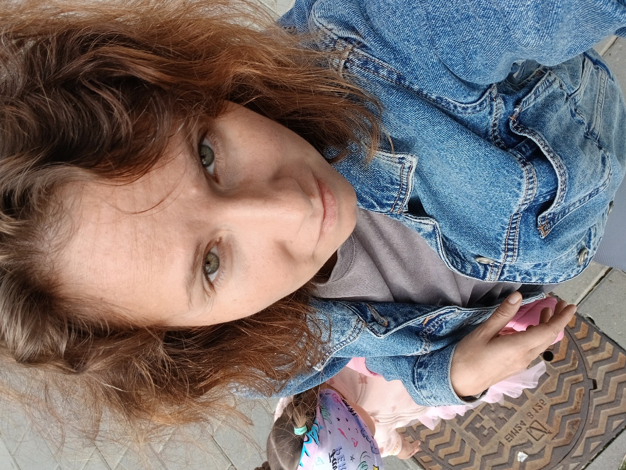 In Yekaterinburg, Russia, a woman with reddish-brown curly hair, wearing a denim jacket over a grey t-shirt, is captured in a high-angle selfie. She has green eyes and fair skin with freckles. Partially visible behind her to the left is a young child wearing a light pink outfit and a colorful fabric mask over their face, their hair in pigtails. They are standing on a textured metal manhole cover, which is common in urban environments. The ground around the manhole cover appears to be paved with grey rectangular stones. The overall setting suggests an outdoor urban location.