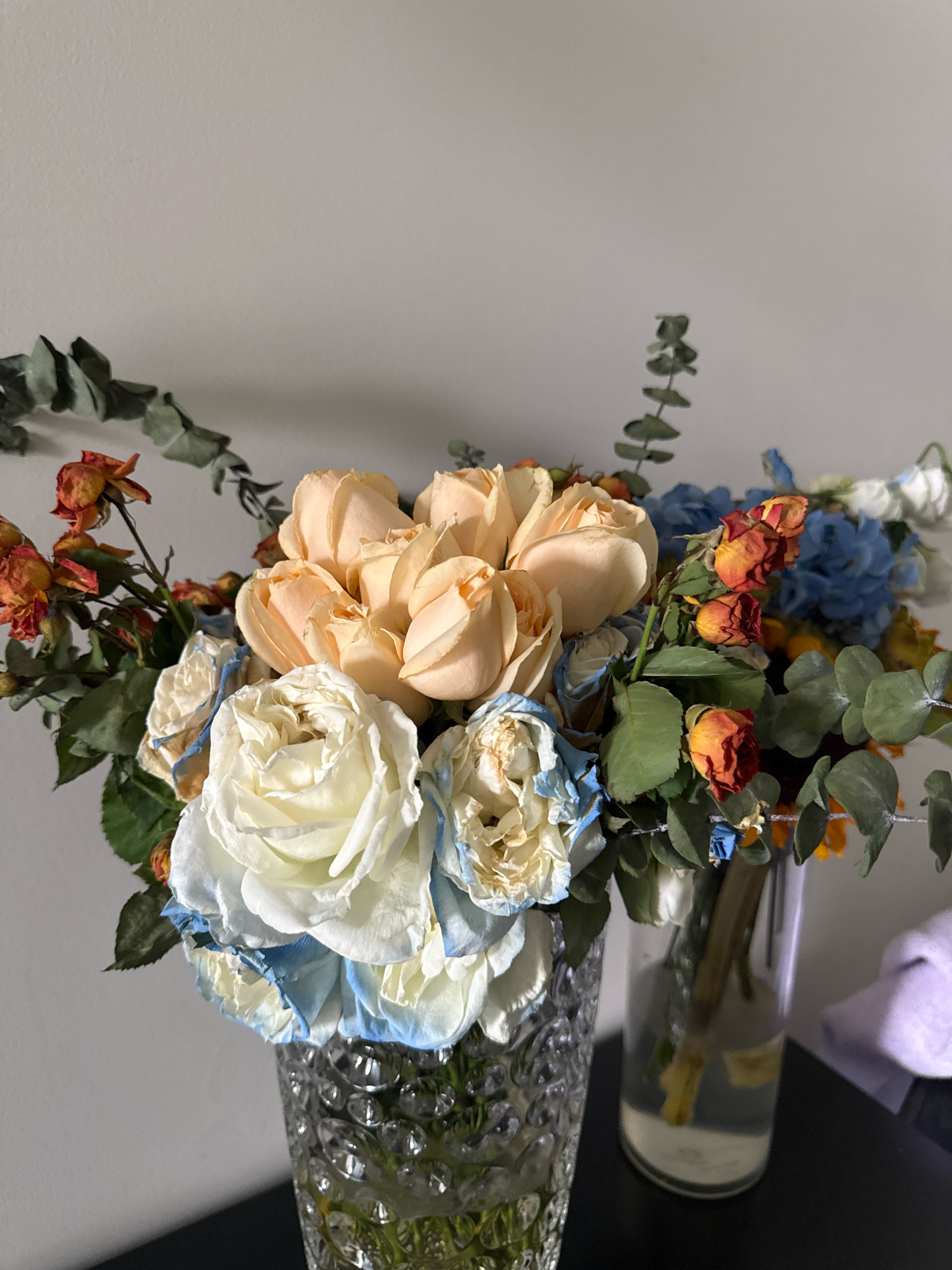 A close-up, eye-level shot shows two vases of flowers on a dark surface in front of a light gray wall. The vase on the left is a clear, patterned glass vase holding a bouquet primarily composed of pale peach roses and white roses with blue-tinted edges. Dried orange flowers and green eucalyptus leaves are interspersed throughout the bouquet. The vase on the right is a taller, clear glass cylinder holding a smaller arrangement with similar flowers, including blue hydrangeas and more dried orange flowers. The lighting suggests an indoor setting, possibly a home or shop, with soft light illuminating the flowers from the left. No people are visible in the image. The scene appears to be located in Yiwu, China, a city known for its wholesale markets, particularly for small commodities and decorative items like flowers.