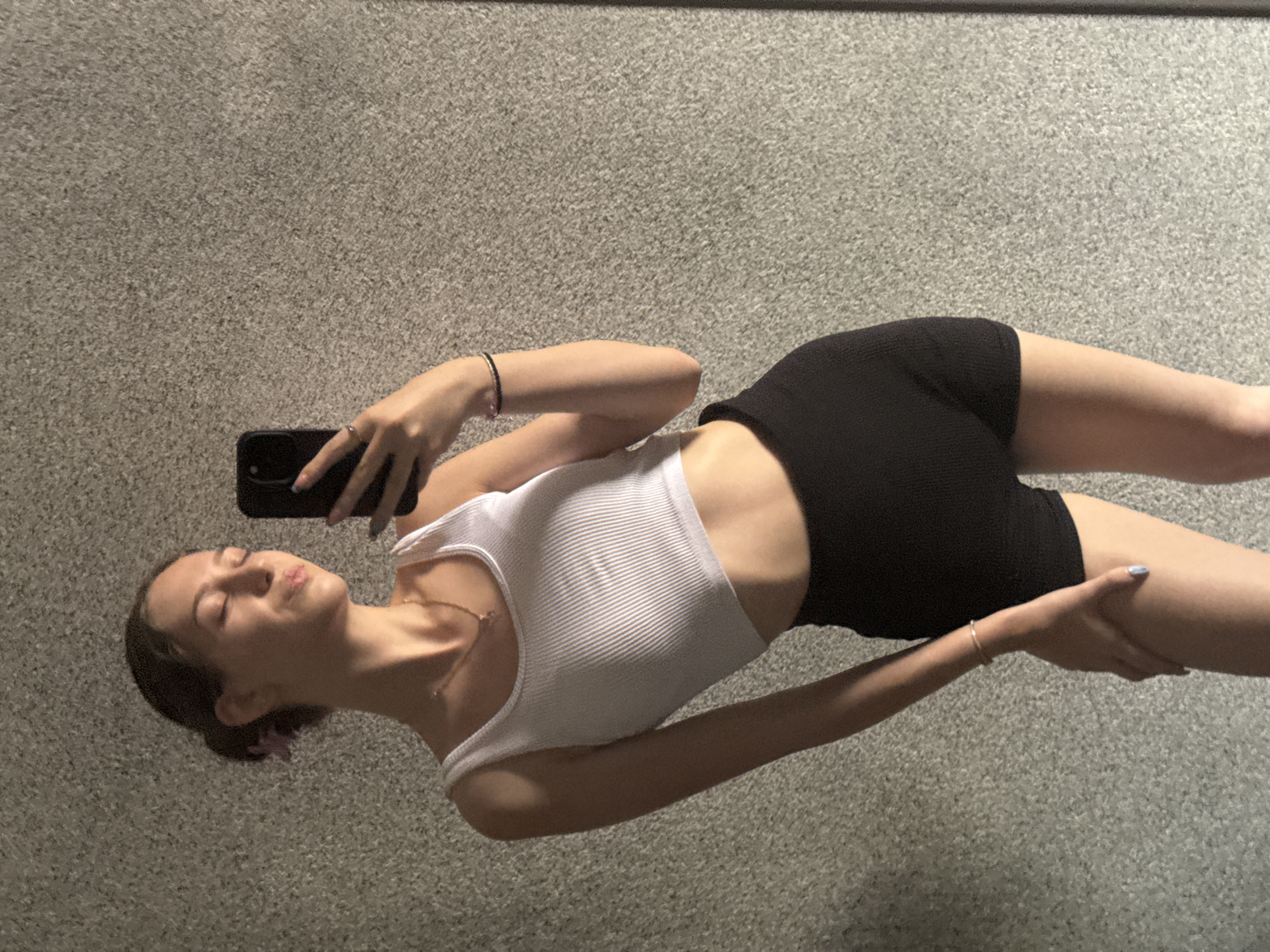A young woman is taking a selfie in front of a textured gray wall. She is wearing a white ribbed tank top and black shorts. Her hair is pulled back in a bun, with a pink accessory. She is holding a black smartphone in her right hand. The lighting suggests it is daytime, with a shadow falling on the wall from the left. The location is not specified in the image or its context.