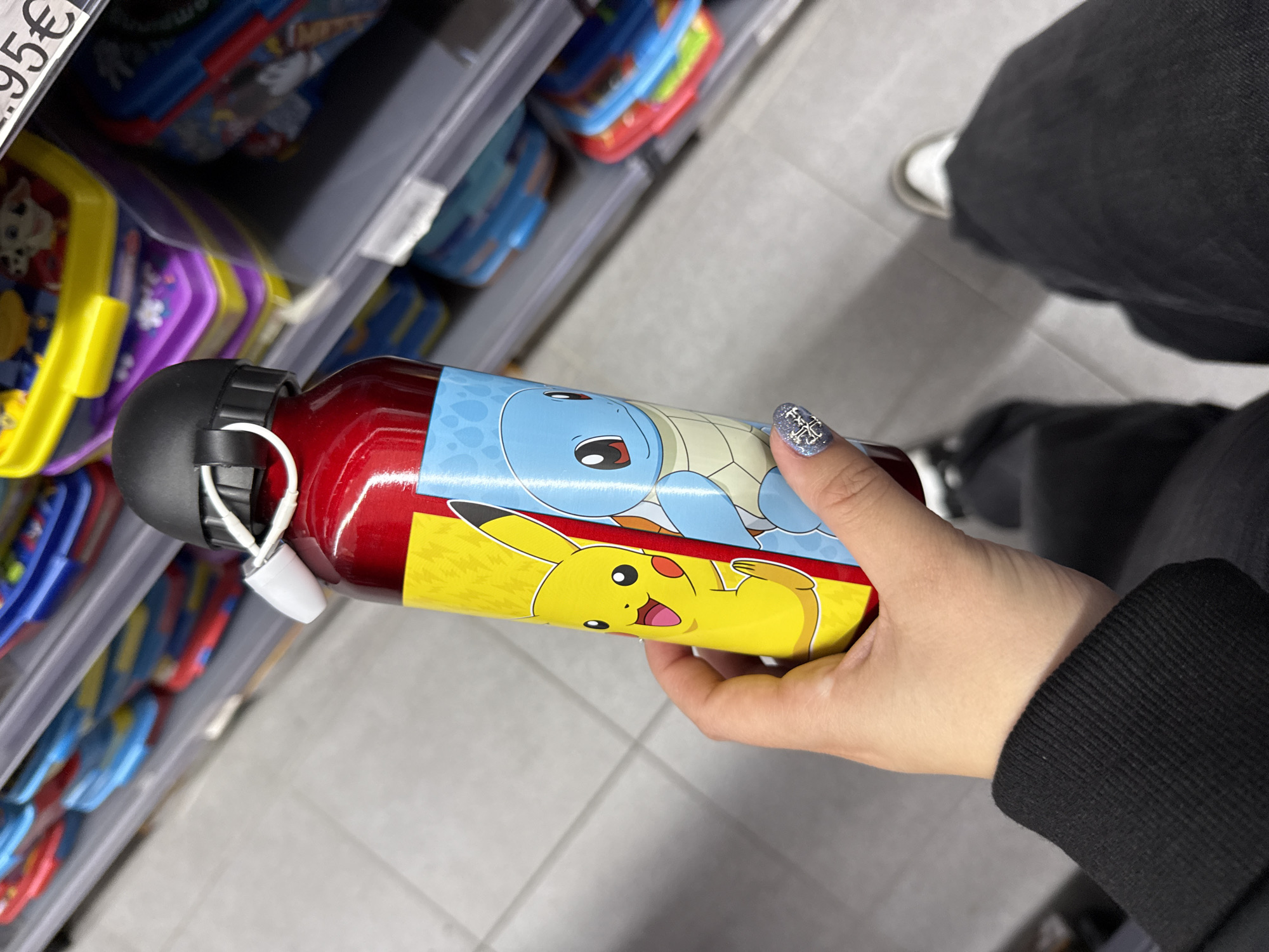 A hand holds a red and yellow water bottle, featuring images of Pikachu and Squirtle from Pokémon, in an aisle of a store. The aisle displays merchandise, including lunch boxes and containers, on gray metal shelves. The person holding the bottle wears black jeans, a black sweater, and white sneakers are visible near their feet. The scene takes place in an indoor setting, likely a retail store, with light-colored tiled flooring. A price tag with "95€" is visible on the left side of the image. The city is not explicitly indicated in the image but the presence of Euro currency implies it is in Europe, and the request specified Madrid, Spain.