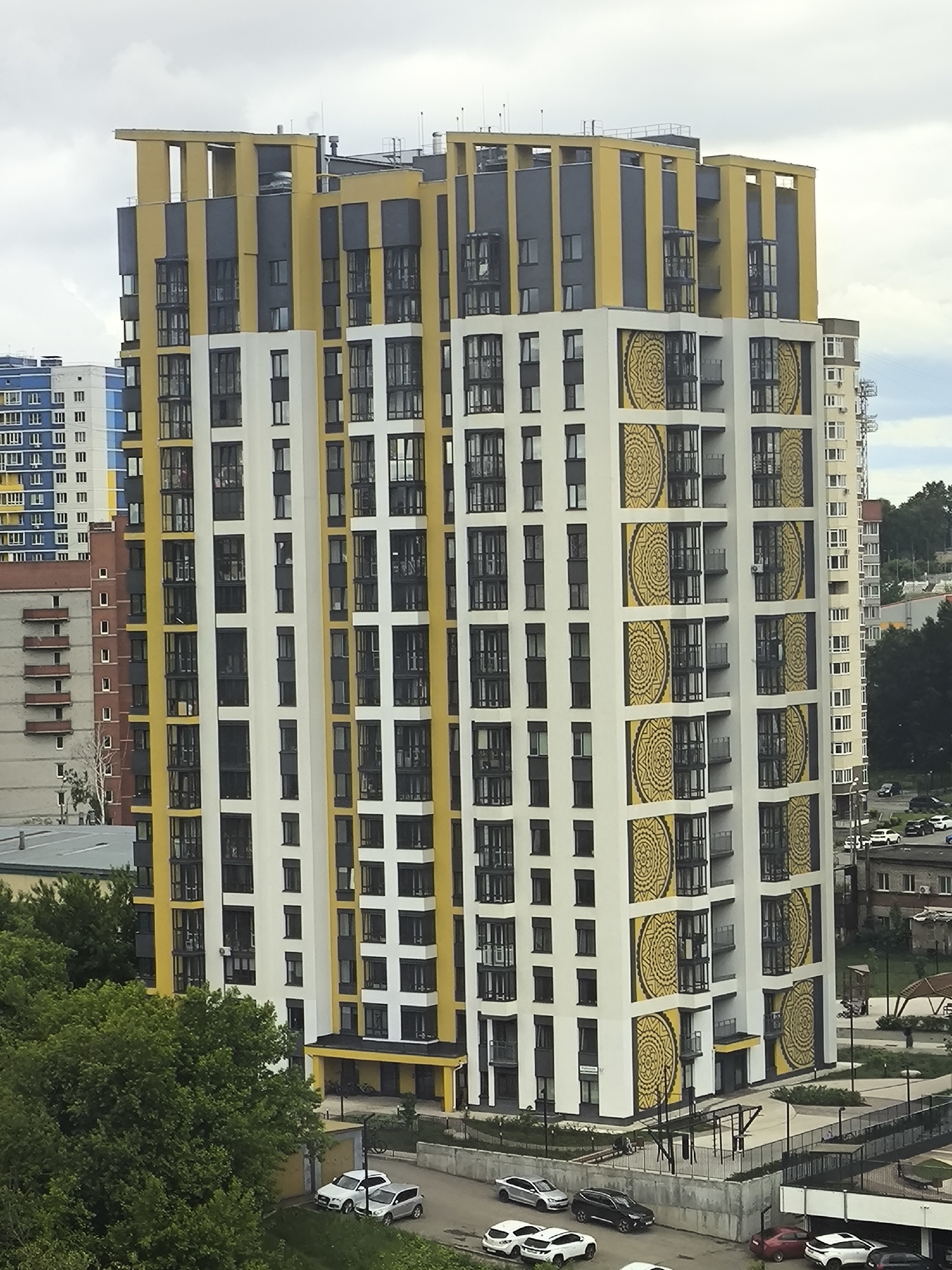 An elevated, wide-shot perspective captures a modern urban residential scene in Unknown, Unknown, under an overcast sky. The central subject is a multi-sectioned, high-rise apartment building characterized by a distinct architectural style. Its facade is segmented into vertical white, yellow, and dark gray panels. The white sections are adorned with large, stacked, circular decorative patterns in yellow and black. The upper portion of the building features prominent yellow structural elements creating a unique crown. Numerous windows with dark frames and some glass-enclosed balconies are visible. To the left and behind the main building, a blue and white high-rise and an older, multi-story concrete building with exposed balconies are present. A lighter-colored high-rise building is partially visible to the right. The ground level features asphalt roads, paved parking areas, and abundant green trees in the foreground and mid-ground. Multiple cars, including sedans and SUVs of various colors (white, silver, black, red), are parked or driving. Concrete retaining walls, fences, and a small outdoor exercise area with metal equipment are also observed. No individuals are distinctly visible.