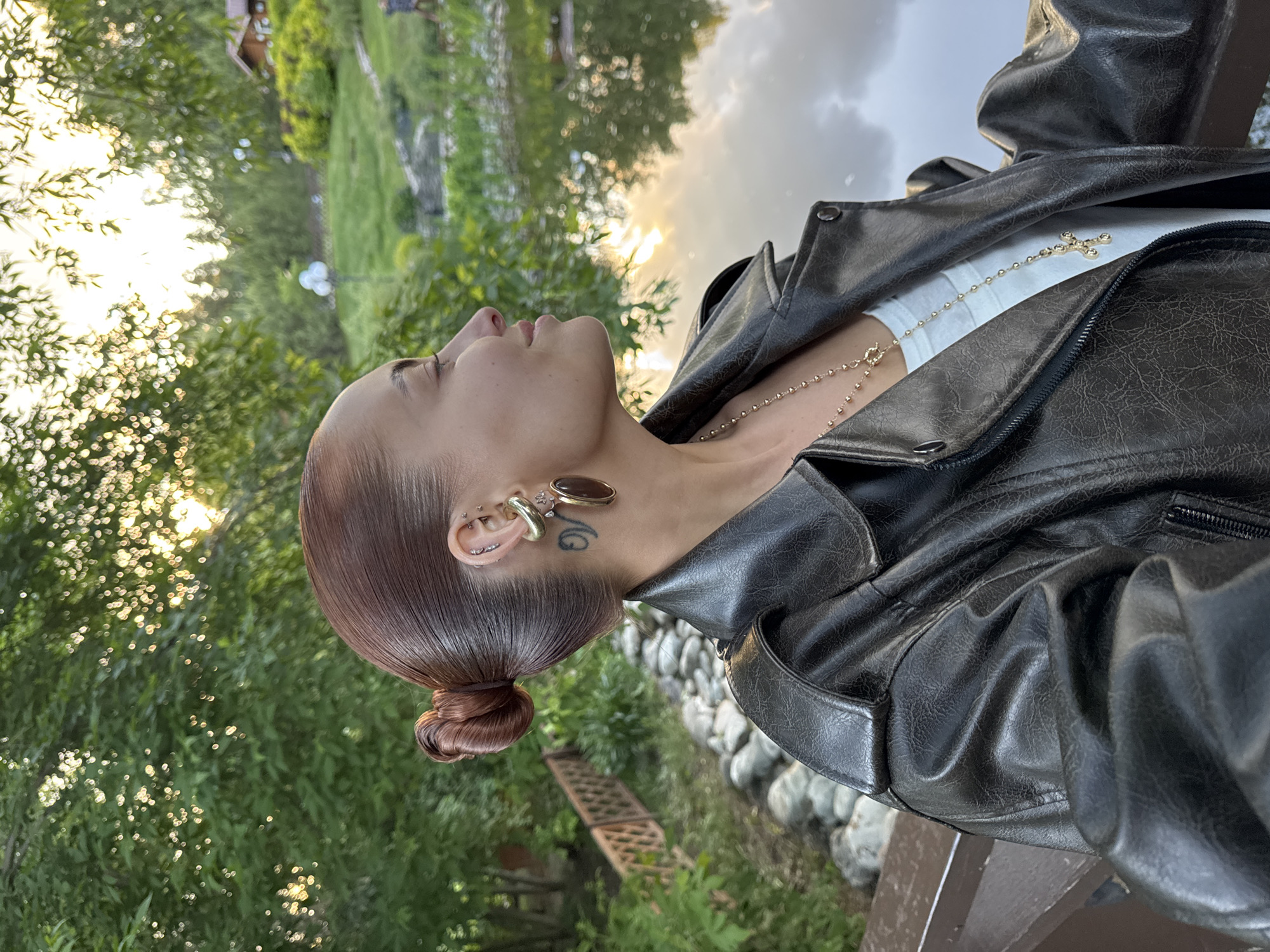A person, likely female, is shown in profile from the chest up, looking upwards towards a bright light source. She has reddish-brown hair styled into a smooth, low bun. Her left ear is adorned with multiple piercings, including studs, a barbell, a gold-toned ear cuff, and a prominent oval-shaped earring. A tattoo of stylized letters is visible on her neck beneath her ear. She is wearing a dark, textured faux leather jacket over a plain white top. A delicate gold chain necklace with a small, ornate cross pendant lies on her chest.
The scene is outdoors, suggesting either sunrise or sunset due to the warm, golden light filtering through dense green foliage in the background, creating a soft bokeh effect. Further back, there are glimpses of grass, scattered rocks, and a rustic wooden structure. The person's gaze is directed upwards, conveying a sense of serenity or contemplation as she poses for the photograph. The jacket's material shows a slightly worn or crinkled texture. The location context is Izhevsk, Russia.