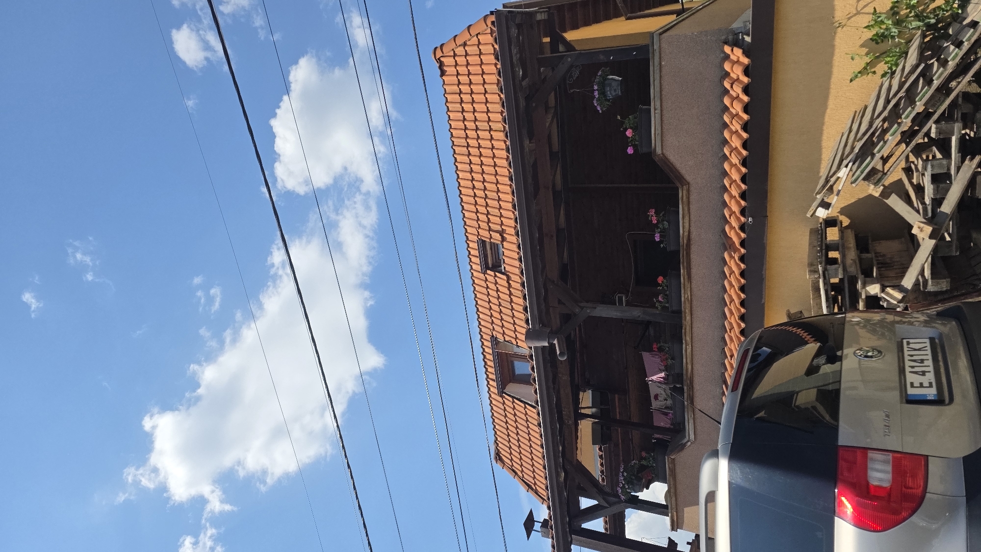 The image shows a building with a red-tiled roof in what appears to be Blagoevgrad, Bulgaria based on the vehicle license plate E 4141 KT. The building has a covered porch or balcony area with flowers. Part of a vehicle is visible in the foreground. There are utility lines in the sky, which is partially cloudy. There is wooden debris and an orange wall. The image is rotated counterclockwise.