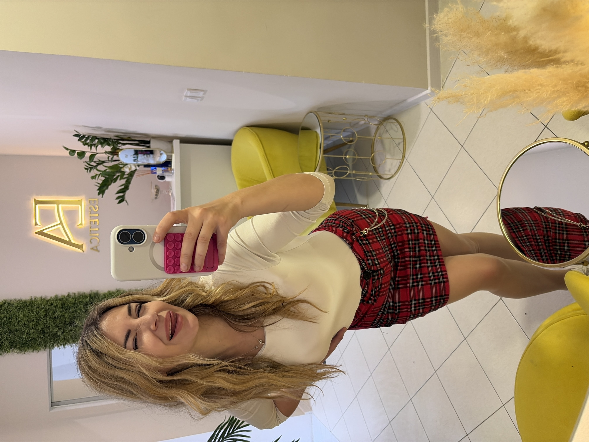 The image is a selfie taken indoors. The woman is fair-skinned with blonde wavy hair, wearing a light-colored long-sleeved shirt and a red plaid skirt. She is holding a phone with a pink grip, capturing her reflection. She's smiling and sticking out her tongue.
The background shows a beauty salon with a lit-up sign "FA ESTETICA," suggesting the location in Budva, Montenegro. There are plants, a yellow chair, a gold decorative cage-like item, a round mirror, and tiled flooring. The lighting is bright, indicating a well-lit interior.