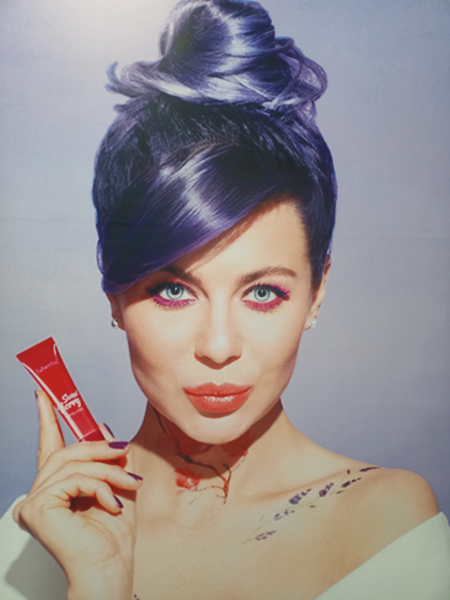 The image is a studio portrait of a woman with purple hair styled in a high bun. She is wearing bright pink eye makeup and red lipstick. She is holding a small red tube of what appears to be makeup in her left hand. There are red and purple floral designs on her neck and shoulder. The background is a plain, light gray. The image does not contain any recognizable landmarks or elements that would indicate its location in Щучинск қаласы әкімдігі, Kazakhstan.