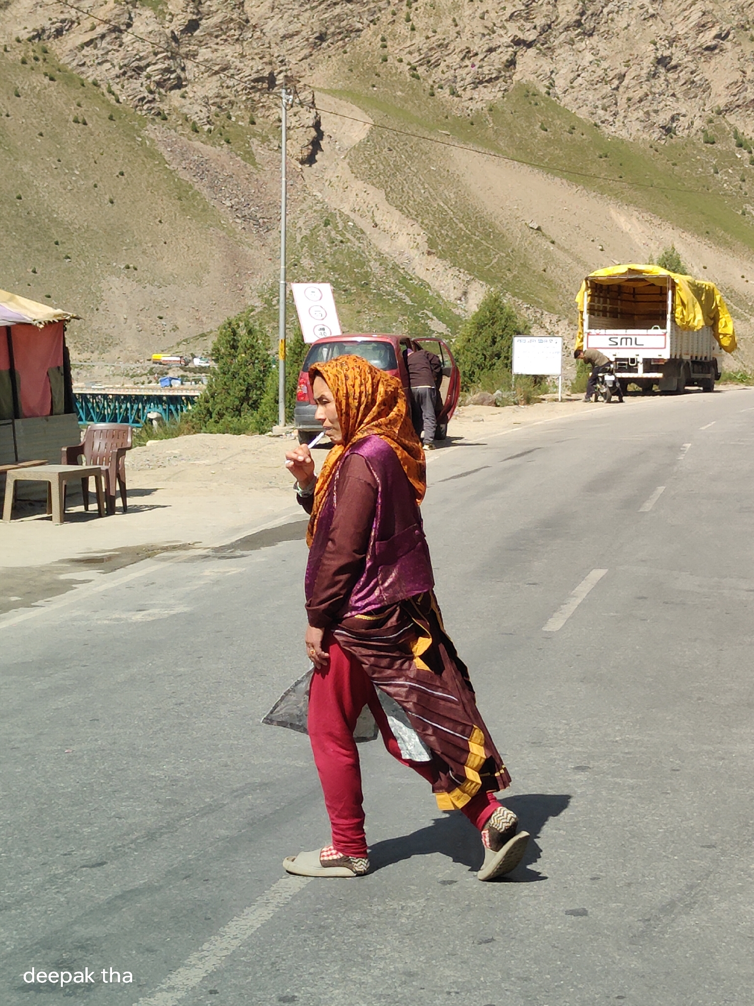 In Darcha, India, a woman wearing a headscarf and a maroon and purple patterned outfit, with red pants and open-toed sandals, walks across a paved road. She is holding a white stick to her mouth. In the background, a red car with its door open is parked on the side of the road. Behind the car, a white truck with a yellow tarp-covered cargo bed is stopped, and a person is leaning over its rear. The road runs through a valley with steep, rocky, and sparsely vegetated hillsides. A street light and a sign with three circles and a number are visible. A temporary shelter with a reddish roof and a table and chairs are situated on the left side of the road.