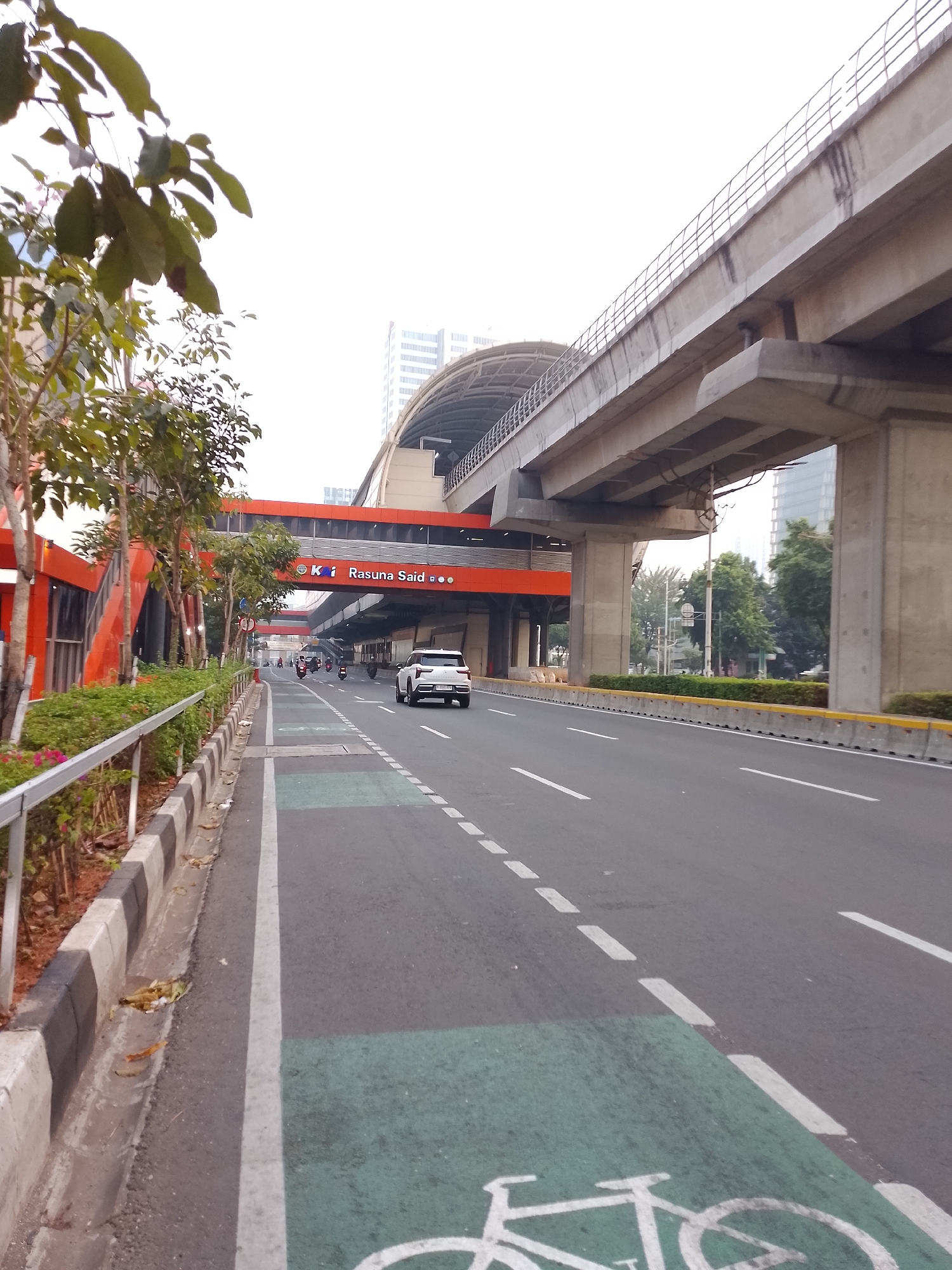 The image depicts a view of the Rasuna Said MRT station in South Jakarta, Indonesia. A white SUV is driving on the road, with several motorcycles also visible. The elevated MRT track and station structure dominate the upper portion of the frame. The street has a designated bicycle lane marked with green paint and a bicycle symbol. Trees and some shrubbery are present along the sidewalk, which is separated from the road by a curb and a metal railing. Buildings can be seen in the background. The sky is overcast.