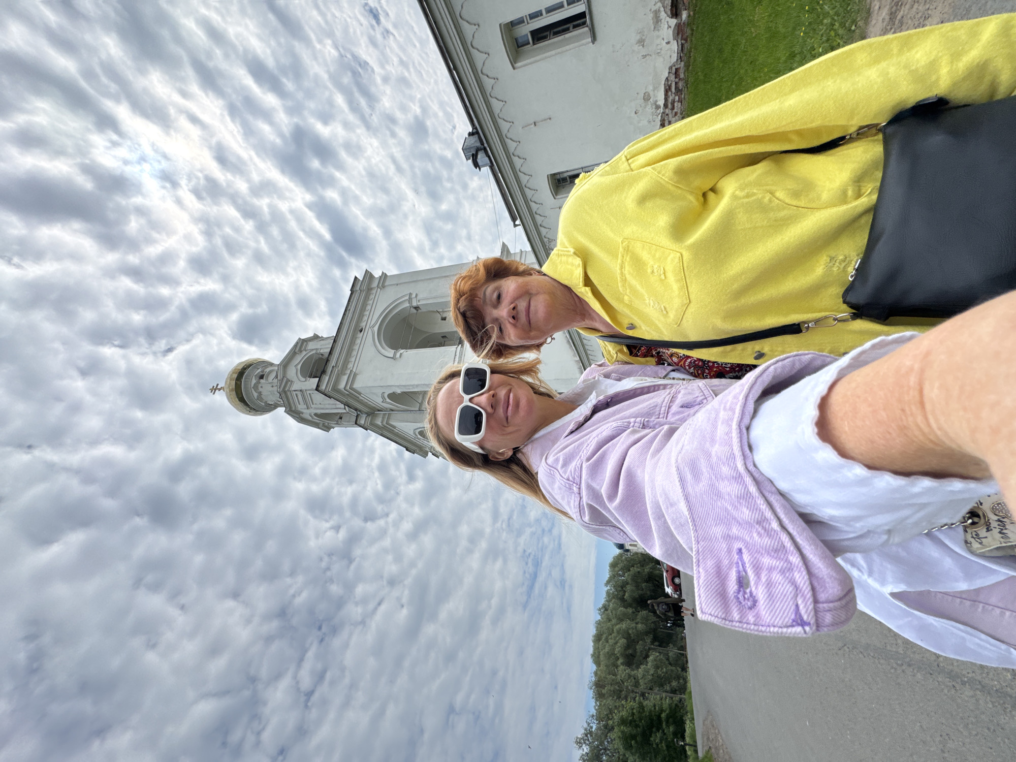 Two women are taking a selfie in front of the bell tower of the Yuriev Monastery in Veliky Novgorod, Russia. The woman in the foreground on the left is wearing large white sunglasses and a lavender denim jacket over a white shirt. The woman on the right is wearing a bright yellow jacket. Behind them, the white bell tower with a golden dome rises against a cloudy sky. To the right of the bell tower is a white building with arched windows, part of the monastery complex. The ground in front of them is an asphalt road, with trees and more of the monastery visible in the background.