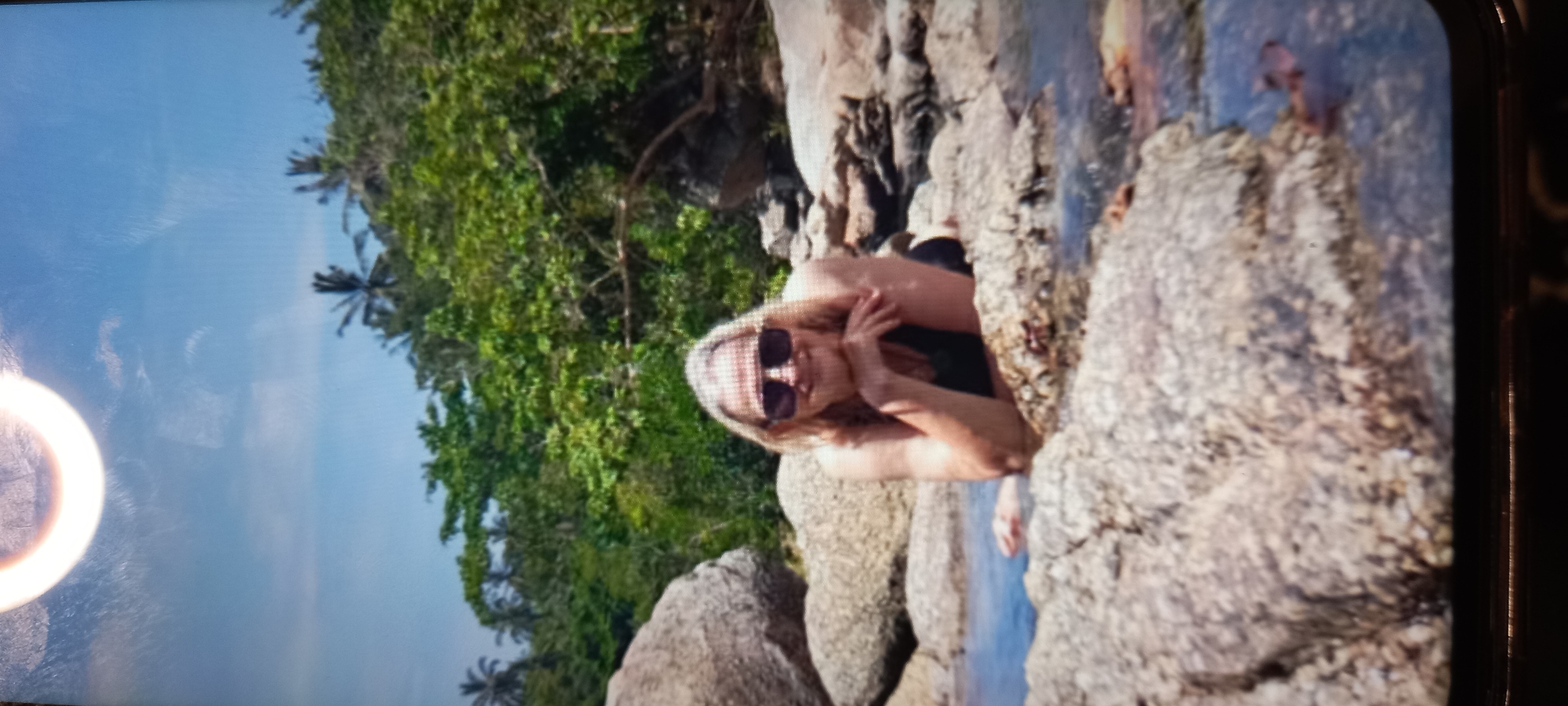 The image, presented sideways, captures a person relaxing in a tropical, rocky environment. A woman with light-colored hair and sunglasses is seen among large, varied-toned rocks, partially submerged in clear blue water. She is wearing a dark top or swimsuit and is looking towards the camera, with her left arm bent and hand near her chest, suggesting a relaxed pose.
In the background, dense, vibrant green tropical foliage, including visible palm trees, rises under a bright blue sky. The setting appears to be a rocky coastline or stream, consistent with the location context of Baan Chaloklum, Thailand. The time of day is bright and sunny, indicated by the clear sky and reflections on the water. A large, circular bright light reflection, possibly glare from a screen or a ring light, is prominent in the upper left of the image. The media appears to be a photograph of a screen, showing some reflections and a partial dark border on the right. No visible text is present.