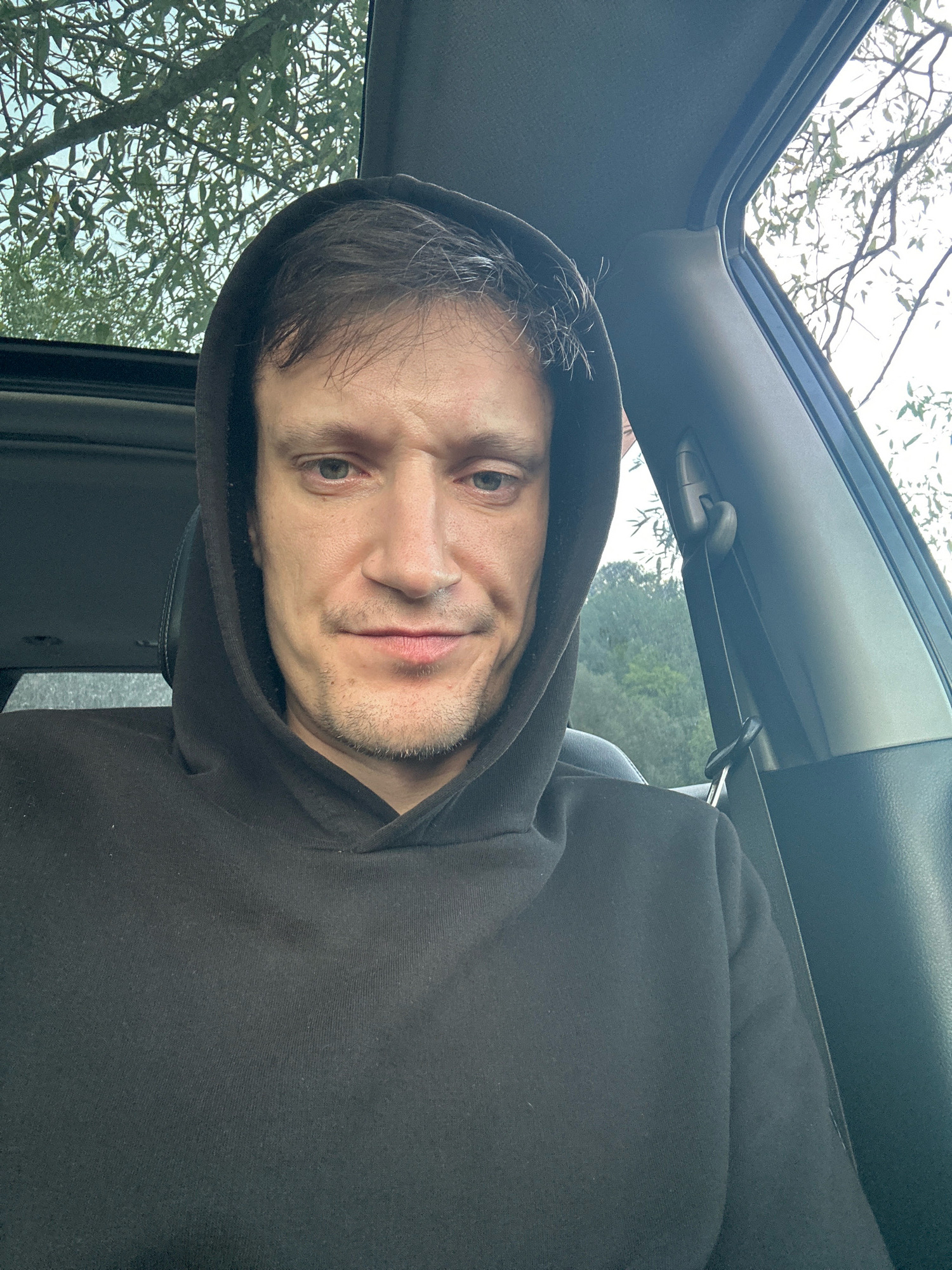 A man wearing a black hoodie sits in the driver's seat of a car. He is looking directly at the camera with a neutral expression. The car's sunroof is open, revealing a view of trees and the sky. The man has short, dark hair and stubble on his face. The interior of the car is visible, including the dashboard and the passenger seat. The image appears to have been taken outdoors, possibly in a rural or suburban area. There is no information about the location in Makiv, Ukraine.