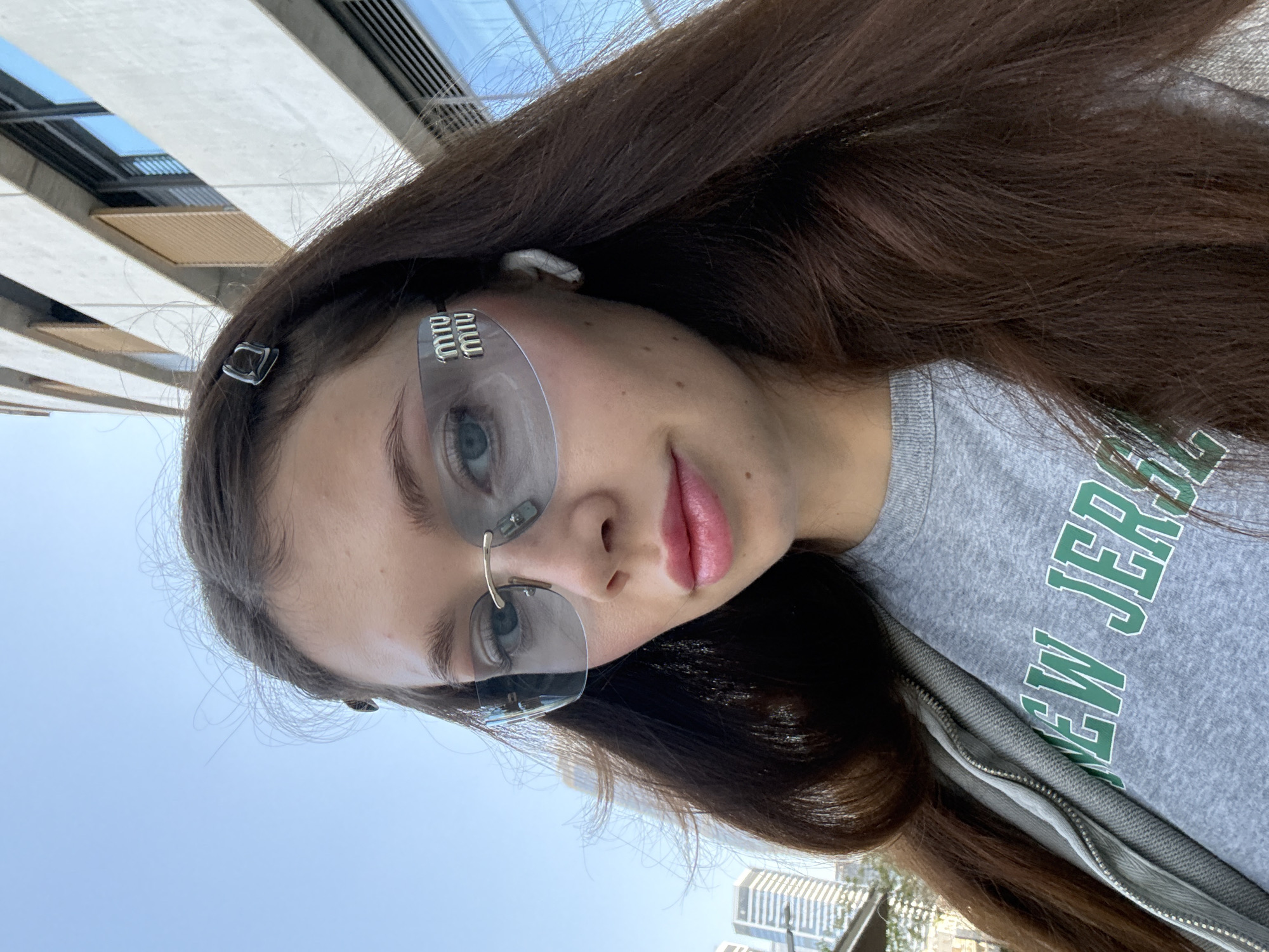The image displays a portrait of a young woman outdoors in an urban setting, likely Tel-Aviv, Israel, on a clear, sunny day. Her face is directed towards the camera, with a neutral to slightly smiling expression and parted lips. She has long, dark brown hair and is wearing stylish, rimless shield sunglasses featuring the "miu miu" logo prominently on the right side (viewer's right) and a gold-toned bridge element on the left side (viewer's left). A small, dark hair clip is visible on her right temple, and a white earbud is partially seen in her right ear. She wears a grey sweatshirt with green block letters spelling "NEW JERS," likely part of "NEW JERSEY," under a grey zippered jacket. The background reveals modern building structures with a clear blue sky, characteristic of urban architecture.