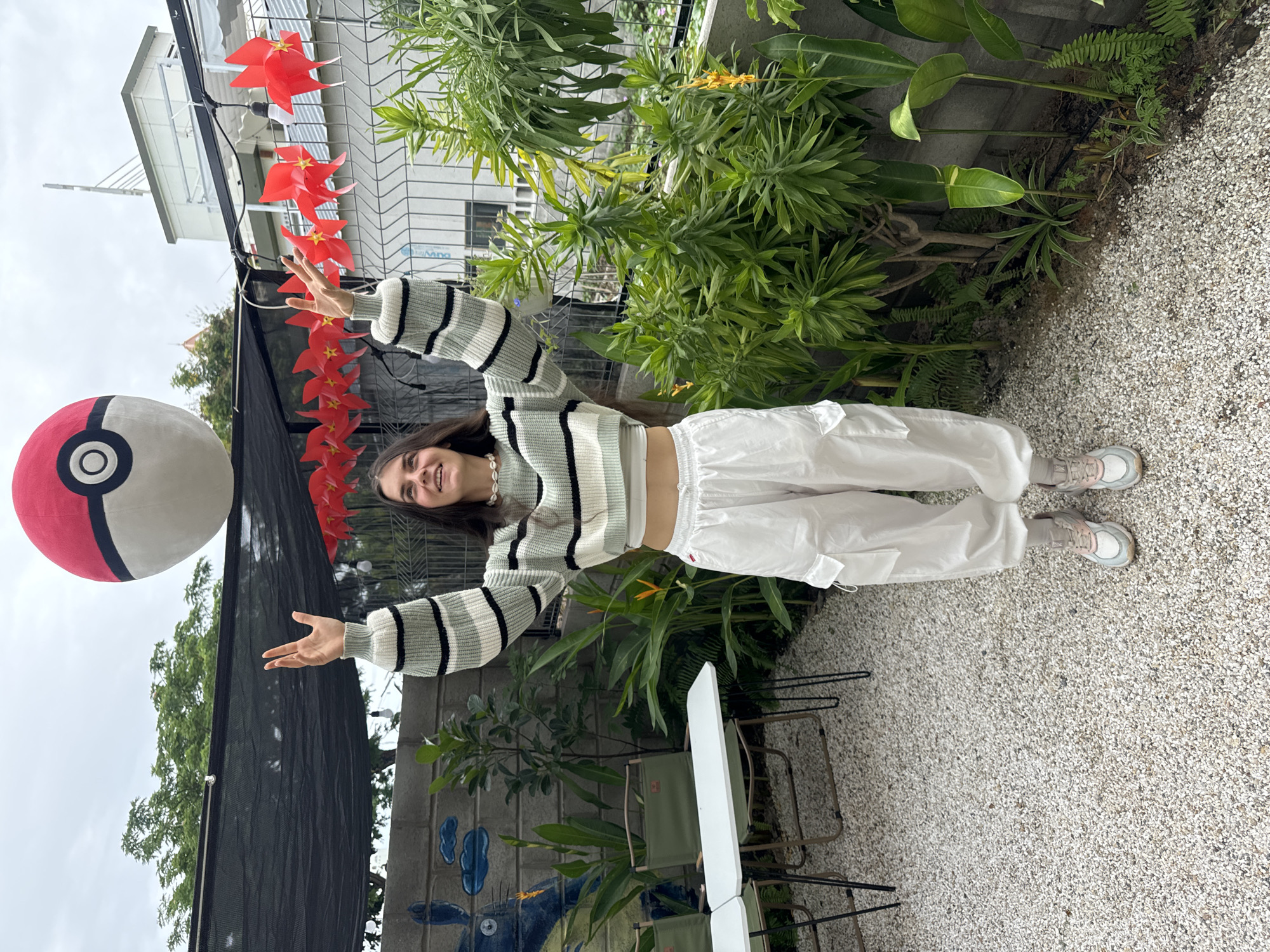 A young woman stands in a courtyard. She has long dark hair and is wearing a light green and white striped sweater, white pants, and pink sneakers. She holds her arms out to either side. A large red and white ball is to her left, partially obscured by a black mesh screen with a string of red pinwheels hanging from it. Behind her, there is a wall with a Totoro mural. Green plants grow along the wall and around the courtyard. The courtyard has a gravel surface and contains tables and chairs. The location appears to be in Phu Thuy Ward, Vietnam. A building is visible in the background. The sky is overcast.