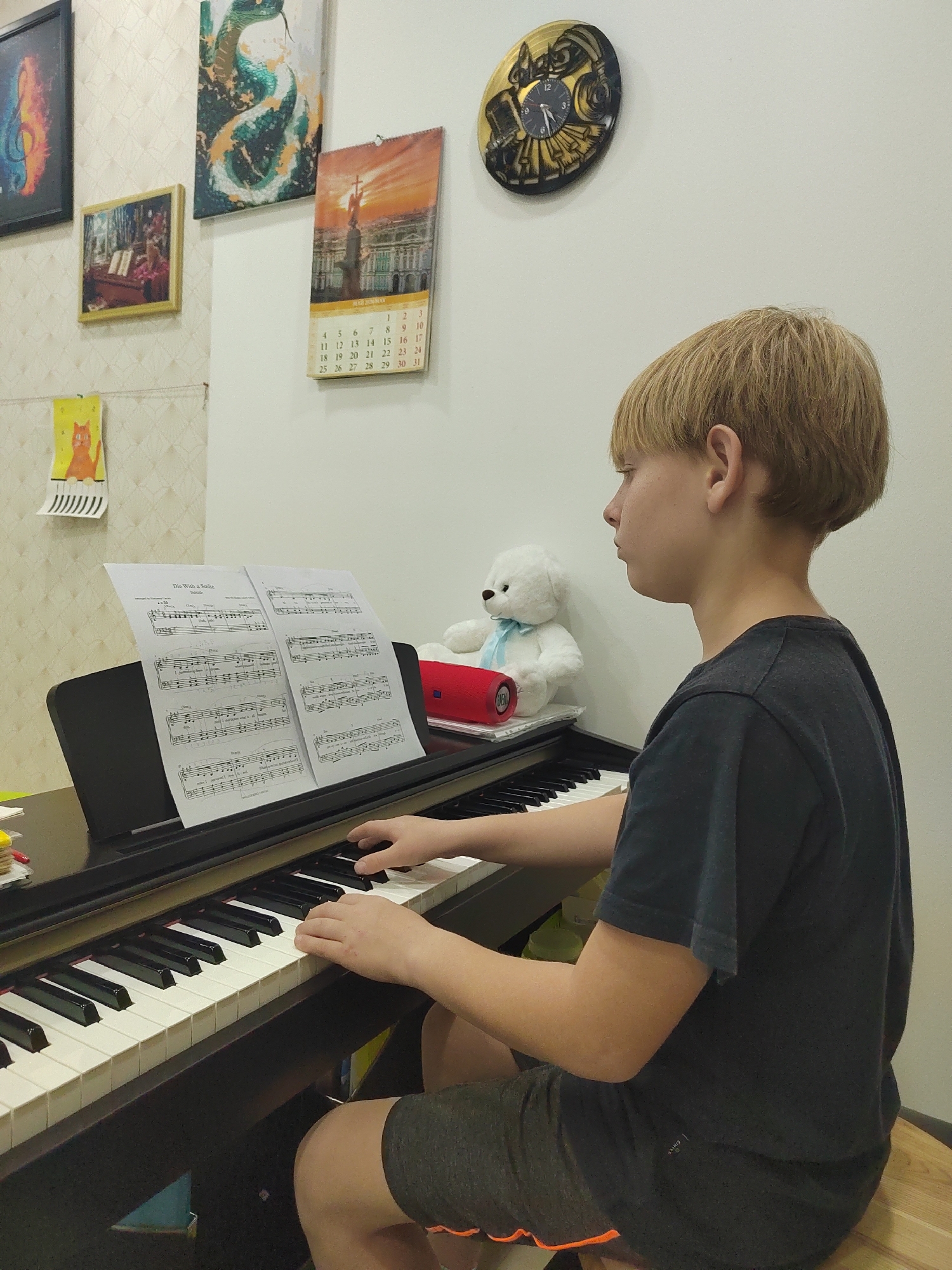 A young boy with blonde hair is playing a black digital piano. He is seated on a wooden bench, wearing a dark gray t-shirt and dark gray shorts. The music sheet placed on the piano reads "The With a Smile". A white teddy bear with a blue ribbon and a red speaker are on the piano behind the music sheet. On the wall behind the piano, there are several framed pictures, a calendar showing the days of the month, and a clock. The wall has light-colored wallpaper, and the room appears to be indoors. The location in the city of Podgorica, Montenegro is undetermined.