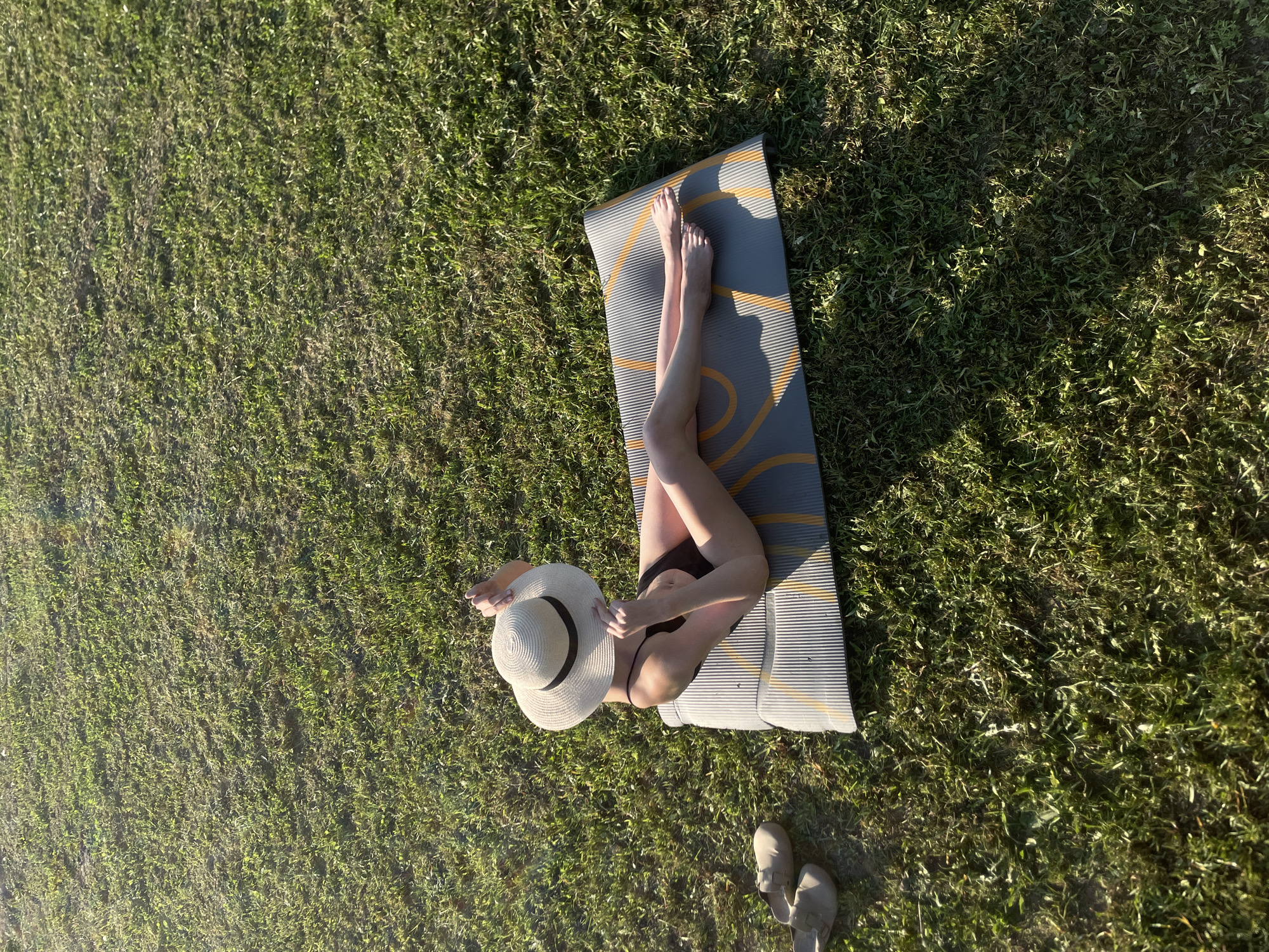 A woman wearing a black bikini and a large straw hat is lying on a gray yoga mat with orange circular designs. The mat is situated on a grassy lawn. The grass is dense and green, suggesting a well-maintained area. A pair of shoes are at the bottom right corner of the image. The scene is in Подшивалово, Russia.