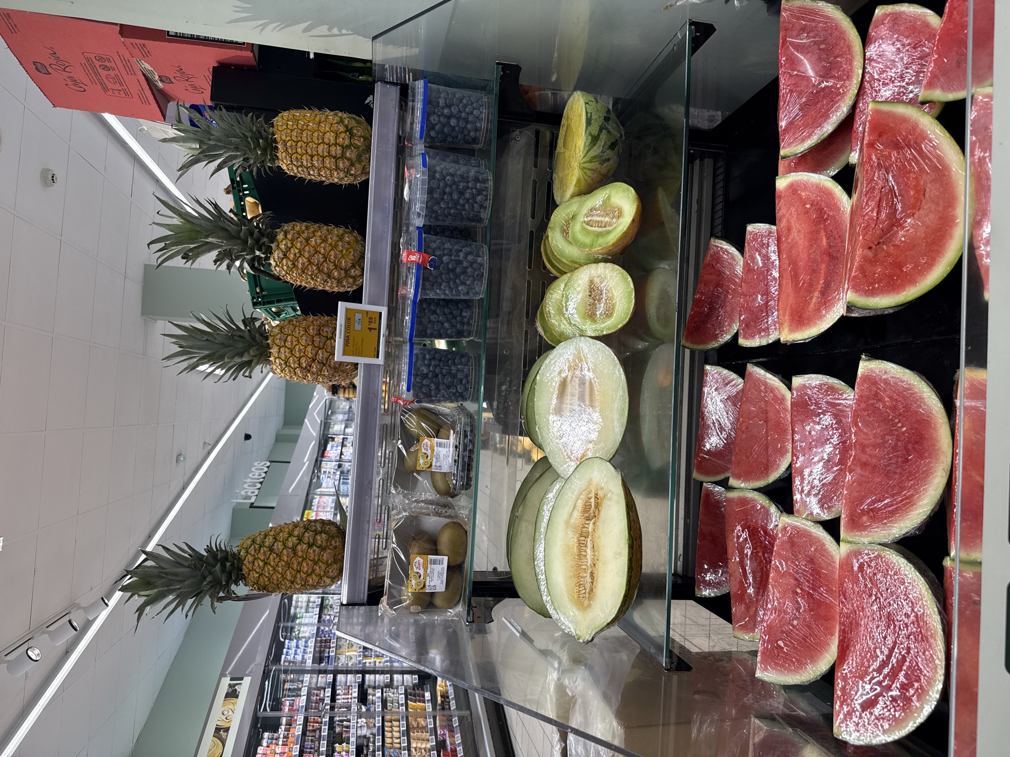 The image shows a display of fruits in a supermarket. In the foreground, several slices of watermelon, wrapped in plastic, are arranged on shelves. Above them are various types of melon, including cantaloupe and honeydew, some of which are also sliced and wrapped. Further up, there are containers of blueberries and packaged kiwi fruit. At the top of the display, a row of pineapples is visible, with one pineapple hanging from above. The background suggests a retail environment with refrigerated cases and shelves stocked with products, along with overhead lighting and signage that reads "Lácteos". The location is specified as Unknown, Unknown.