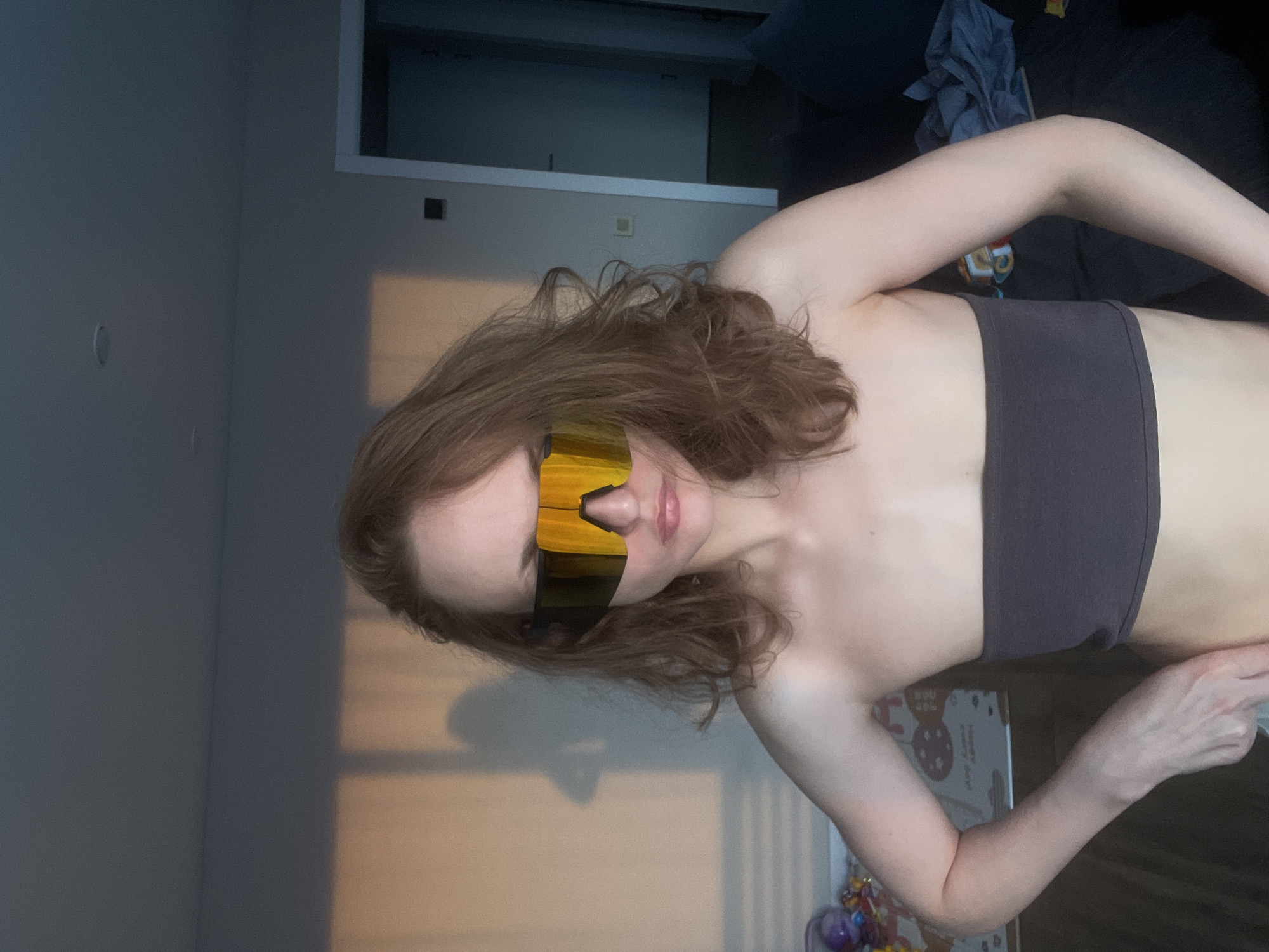This portrait photograph, taken in Podshivalovo, Russia, features a young woman posing indoors. She is captured from the waist up, with her body angled slightly. Her long, wavy brown hair cascades around her shoulders. Her eyes are obscured by a pair of large, futuristic-style mirrored sunglasses with yellow and orange gradient lenses. She is wearing a simple dark grey or black strapless bandeau top, revealing her shoulders and midriff.
The setting appears to be a room, likely a bedroom or living room, with light-colored walls. A doorframe is visible in the upper left corner of the background. Natural light enters from an unseen window, casting a warm, rectangular shadow on the wall, suggesting it may be late afternoon. In the lower right, behind the woman's arm, part of a white box is visible, adorned with cartoon rabbits or bears and the text "HAPPY DAY." Colorful, round objects, possibly ornaments or toys, are also partially visible nearby. The woman's expression, though partially hidden by the sunglasses, appears calm with slightly parted lips, indicating a relaxed and casual pose for the photograph.