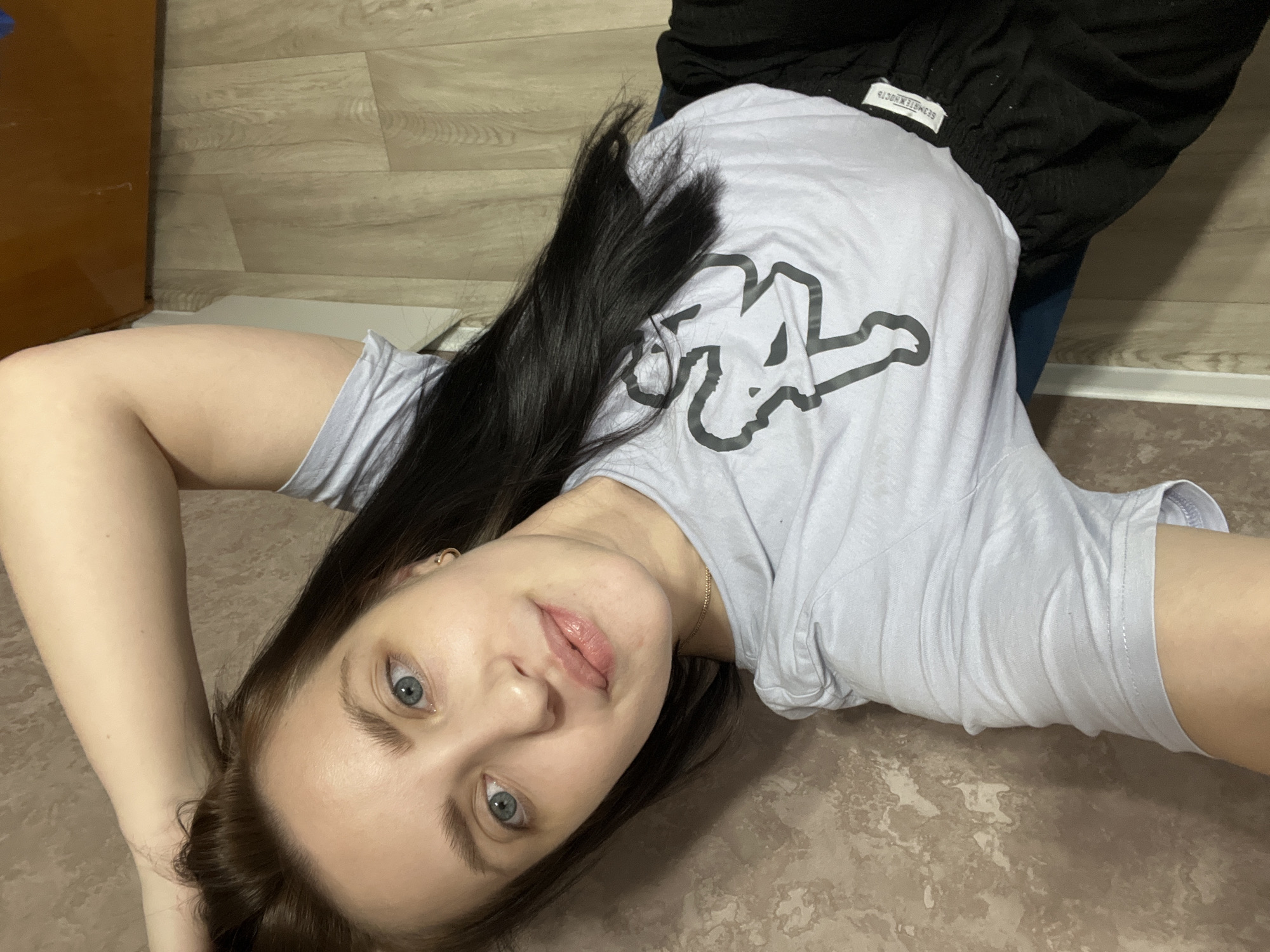A woman with blue eyes and long black hair is lying on a marble floor. She is wearing a light grey Kappa T-shirt and black sweatpants. The floor is beige with a marble pattern. The location is Марковский, Russia.