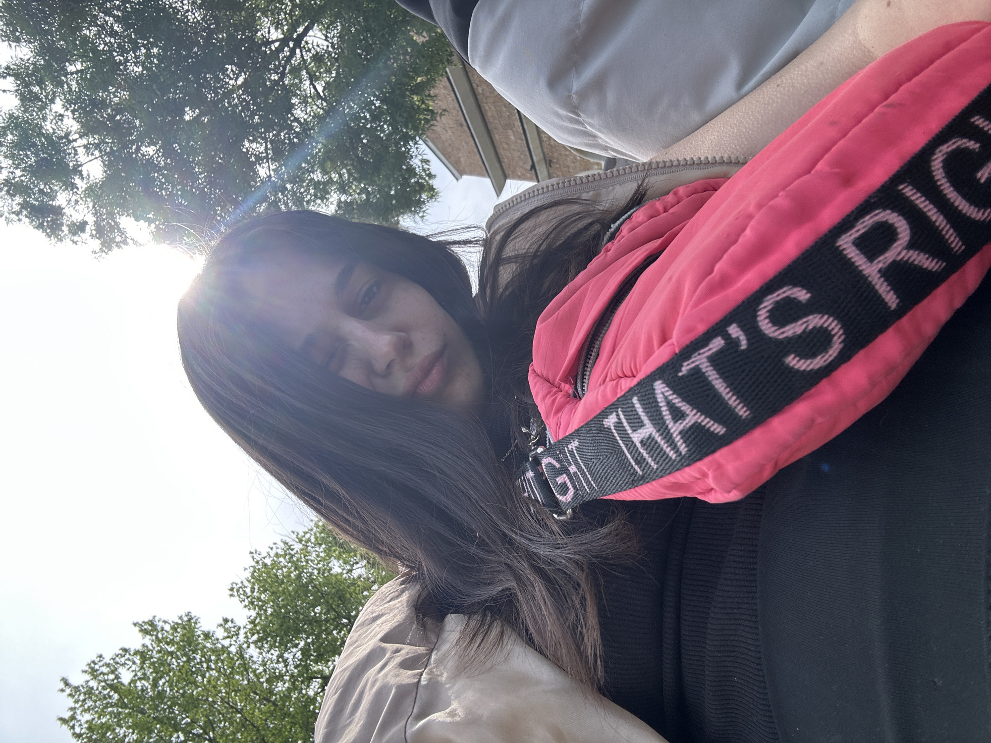 A young woman is photographed from a low angle, possibly a selfie, in an outdoor environment. The location appears to be within a city, indicated by buildings and trees in the background. The photograph is taken during daylight hours, with the sun visible, creating a lens flare. The woman has long, dark hair. She is wearing a beige jacket over a black top. A pink padded accessory or garment with the text "THAT'S RIGHT" is prominently positioned in the foreground. The trees in the background appear to be deciduous, and the sky is visible in the upper portion of the frame. The location is not identifiable as Veliko Tarnovo, Bulgaria, based on the visual information.