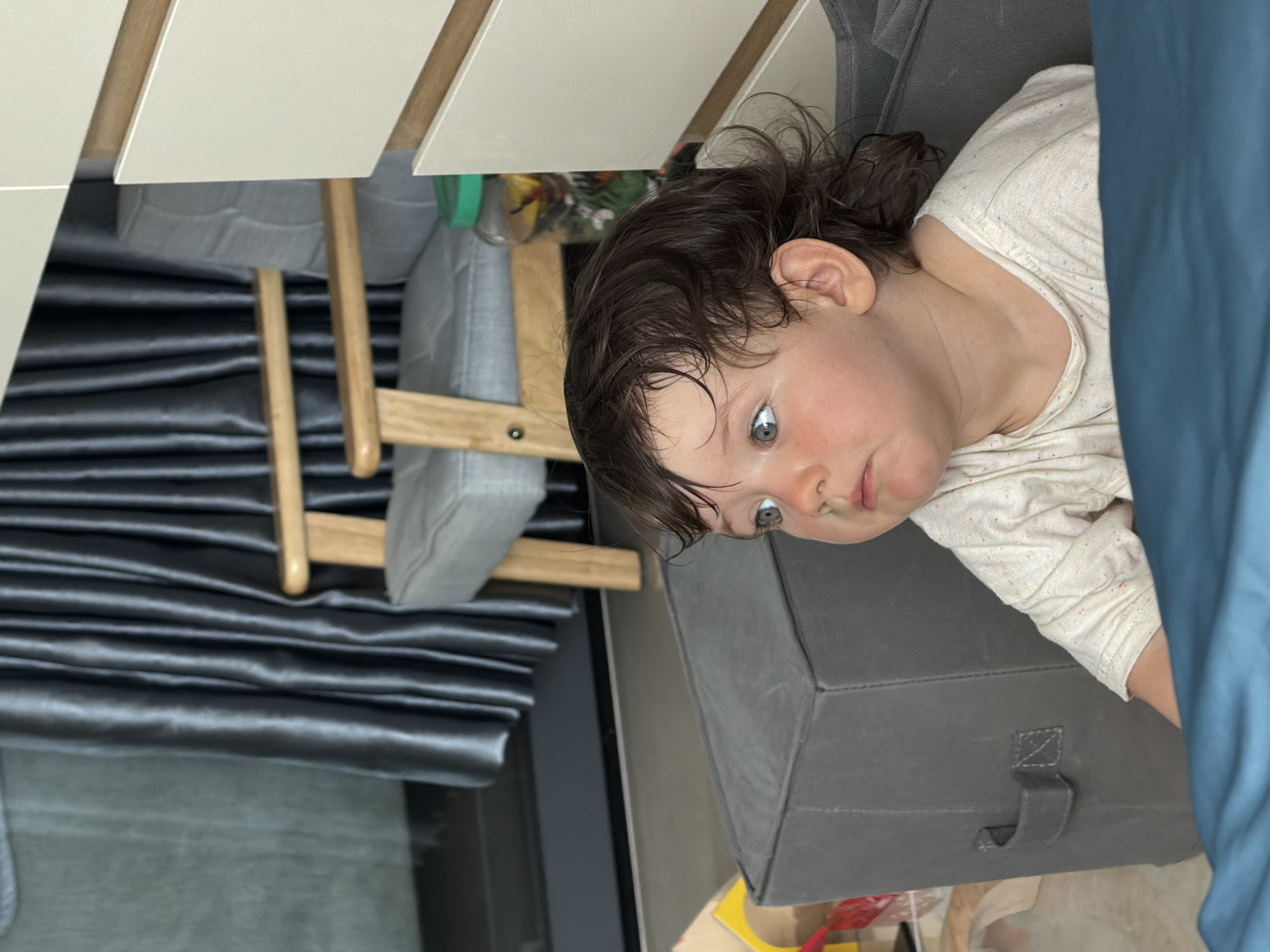 A toddler with dark, curly hair and blue eyes gazes to the right of the frame, positioned in the foreground of a domestic setting. The toddler is wearing a light-colored, patterned t-shirt. Behind the child, a grey cube-shaped ottoman with a handle is visible. Further back, a grey armchair with wooden armrests and legs is present, juxtaposed against dark grey curtains that hang from a window or door. To the right of the armchair, a portion of a white cabinet with wooden trim is visible. The environment appears to be indoors, with the lighting suggesting daytime. No specific city or country details are discernible from the visual information provided.