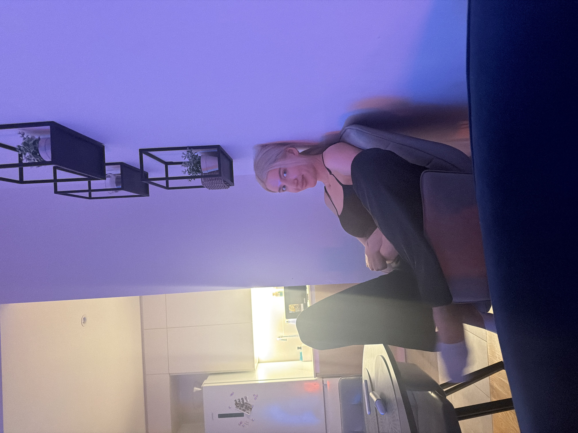 A young woman with blonde hair is sitting in a chair in an apartment. She's wearing a black tank top and dark pants. The apartment has white walls, a white refrigerator, and shelving units with plants. There is a kitchen visible. The location is potentially Belgrade, Serbia.