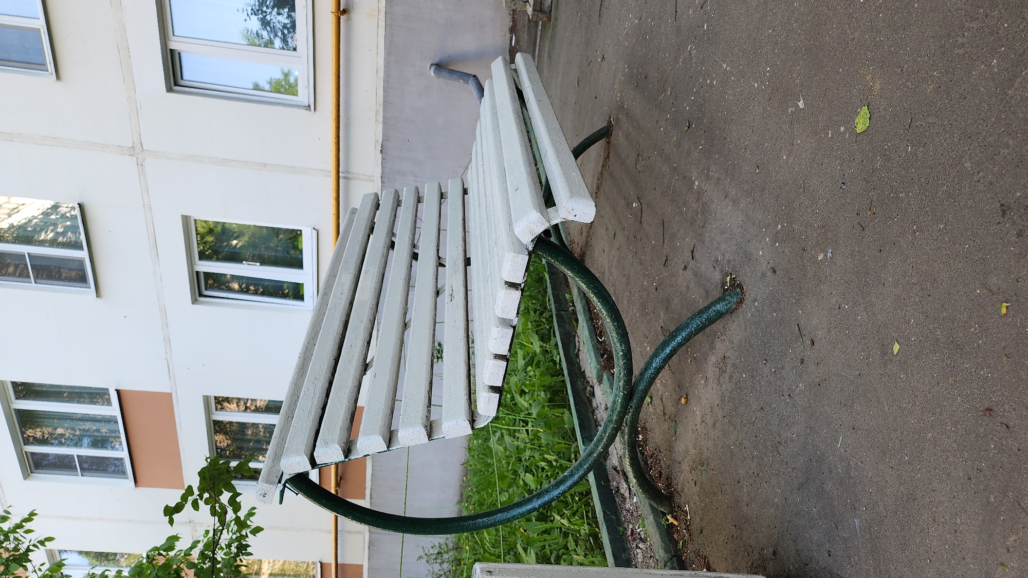 A park bench sits on a paved surface in Moscow, Russia. The bench has a white, slatted seat and a green, curved metal frame. It is positioned near a building with white walls and several windows. Grass and vegetation are visible around the bench. The paved surface appears to be a sidewalk or small plaza.