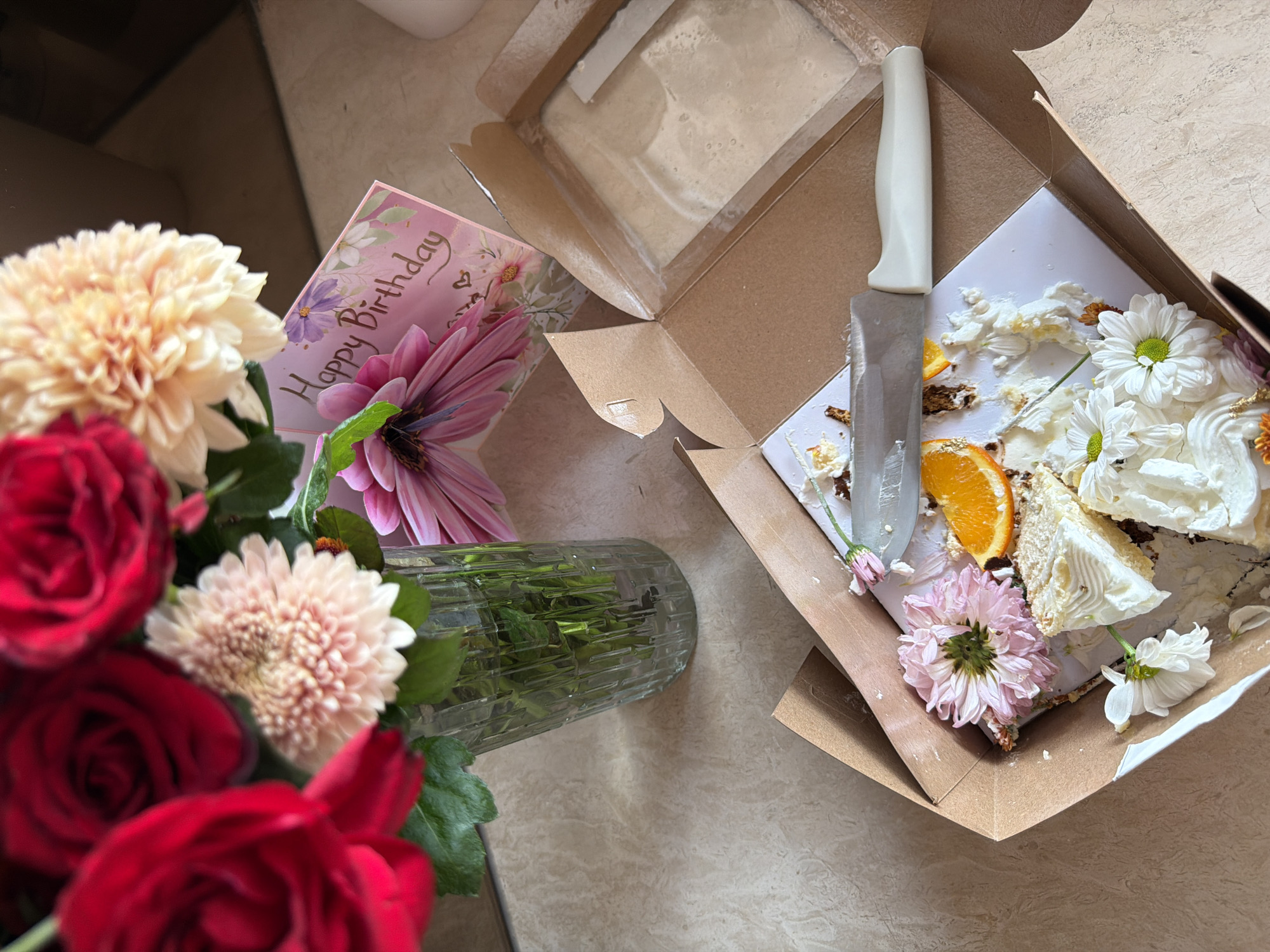 A top-down view shows a still life arrangement on a beige surface. A bouquet of flowers, featuring red roses and pale pink chrysanthemums, sits in a clear, vertically ribbed vase on the left. Next to the flowers is a pink "Happy Birthday" card. A partially eaten cake is in an open cardboard box. A knife with a white handle rests on the cake. The cake appears to have been decorated with white frosting, orange slices, and white and pink flowers. The top of a second box sits behind the other box. The scene does not appear to be in Mengwi, Indonesia.