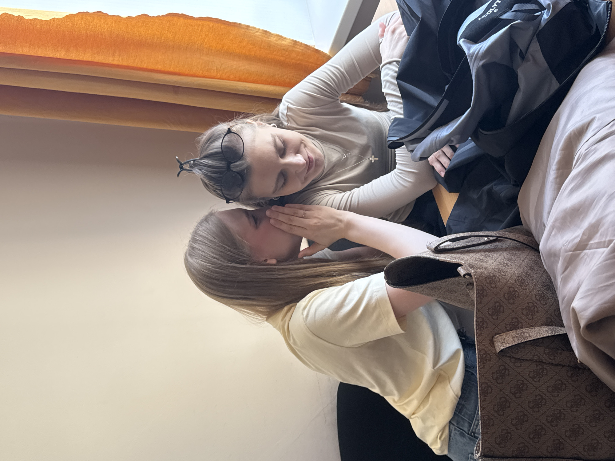 The image shows two young women engaged in a private, intimate conversation indoors, likely in Perm, Russia. The photograph is taken from a slightly elevated, tilted perspective.
One woman, positioned at the upper part of the frame, has blonde hair, with black-rimmed glasses resting on her head. She wears a light-colored long-sleeved top and a necklace adorned with a cross pendant. Her eyes appear closed or cast downwards, and she displays a soft, attentive expression as she listens intently.
Leaning in towards her is another young woman, also with blonde hair, dressed in a light yellow short-sleeved top. She is captured in the act of whispering into the first woman's ear, with her left hand gently covering her mouth in a gesture of secrecy. Her right hand is near the other woman's ear, reinforcing the intimate nature of their interaction. This suggests a moment of shared confidence or a secret being exchanged.
Around the individuals, a few objects are visible. A black jacket or bag, bearing the partial text "ACOL" (possibly "ACOLYTE"), is seen behind the listening woman. To the right, a patterned brown tote bag, featuring a repeating logo reminiscent of the Guess brand, is partially visible. A light beige or brown blanket or duvet occupies the far right of the frame. The background features a plain light-colored wall and an orange curtain or blind at the top, indicating an indoor setting, likely bathed in natural daylight. The overall emotion conveyed is one of trust, closeness, and confidentiality between the two individuals.