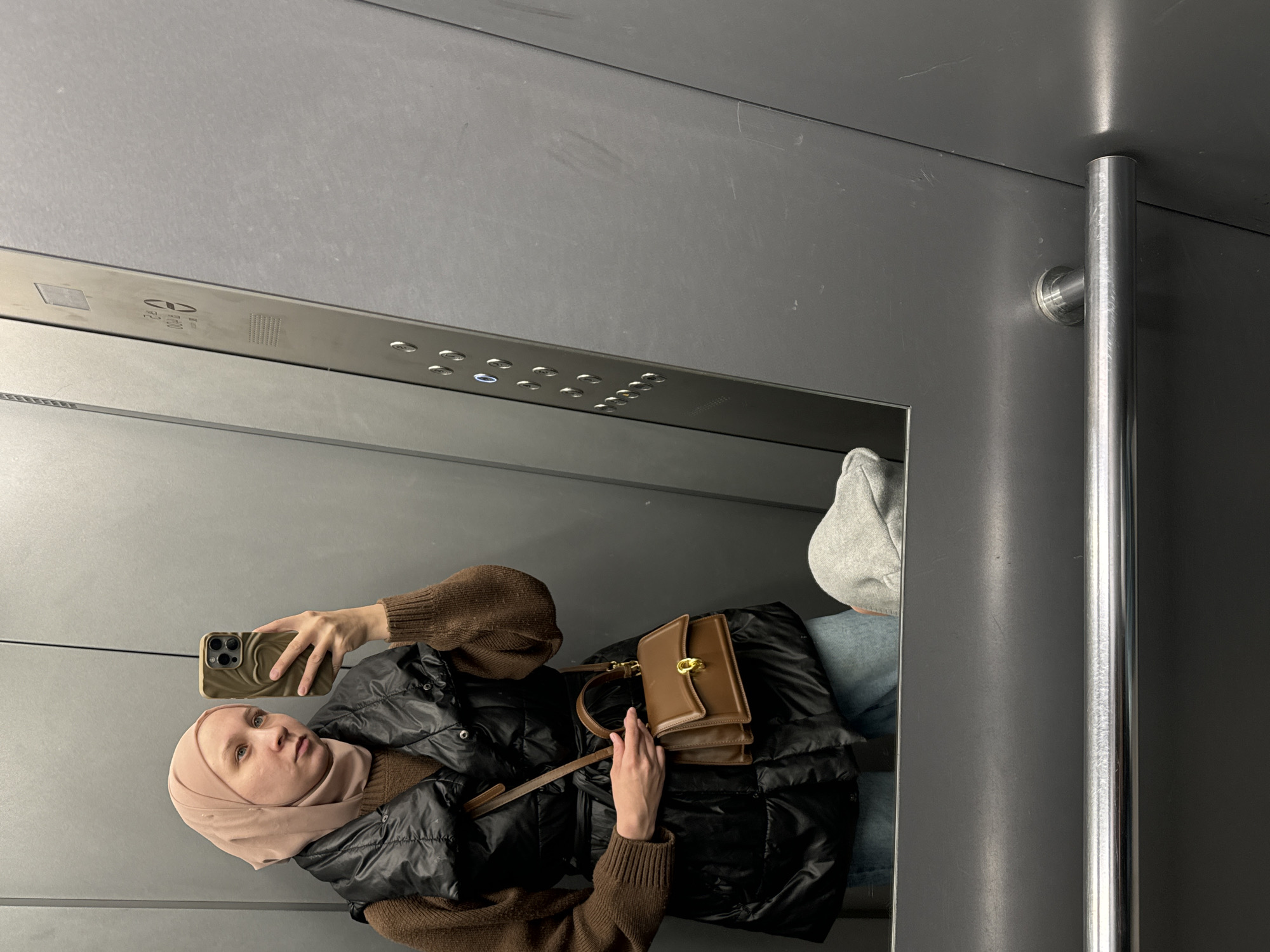 A woman wearing a hijab, brown sweater, and black puffer vest is taking a selfie in an elevator. She is holding her smartphone in her left hand, and her right hand is near a brown purse with a strap. The elevator has stainless steel walls and a control panel with buttons. A silver handrail is visible on the right side of the frame. There is no indication of the location being Chaikovsky, Russia.