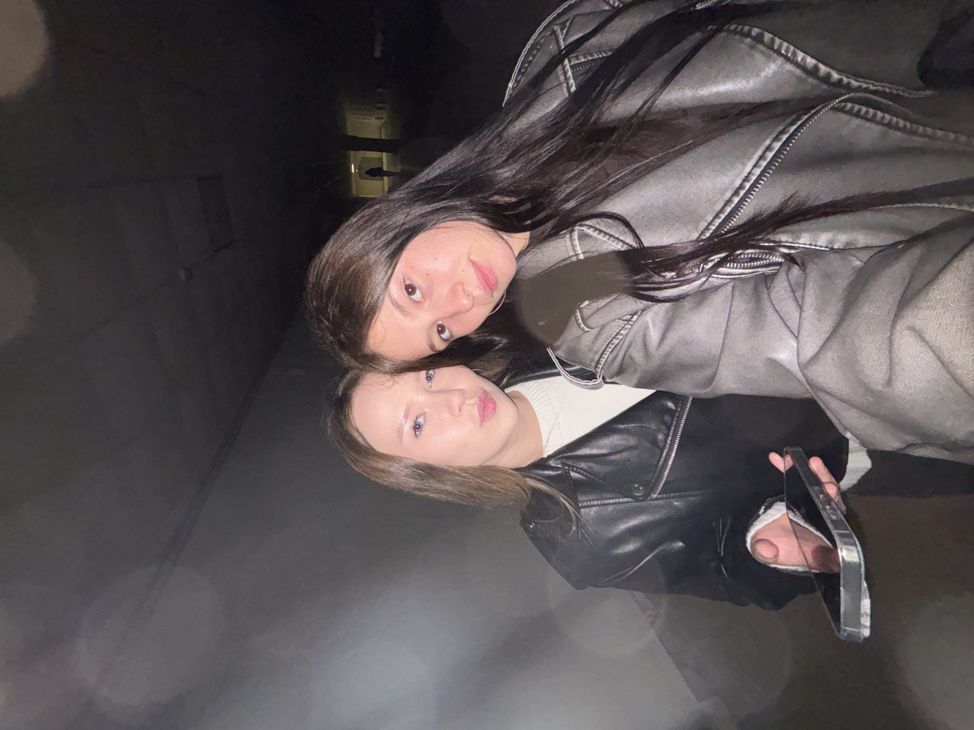 Two young women pose for a selfie in what appears to be an underpass or tunnel. The woman on the left has blonde hair and is wearing a black leather jacket over a white sweater. She is holding a smartphone in her left hand, which is angled to capture the selfie. The woman on the right has long dark hair and is wearing a gray leather jacket. The environment is dimly lit with visible concrete walls and a ceiling that appears to be constructed of panels. In the background, a faint silhouette of a person can be seen at the far end of the tunnel. The overall lighting suggests it is nighttime or a poorly lit indoor space. The location is identified as Chaikovsky, Russia.