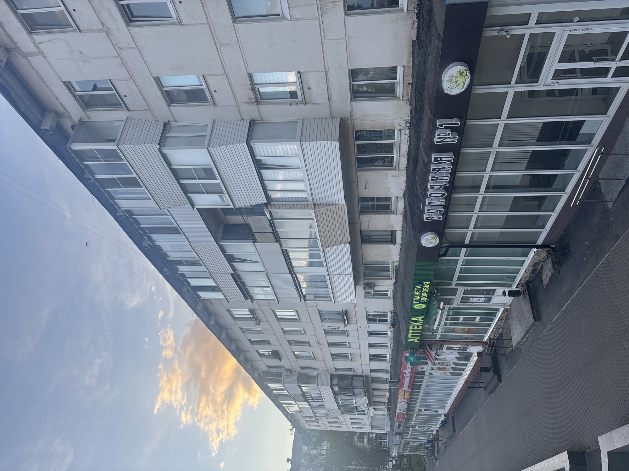 The image shows an urban street in Chaikovsky, Russia, with a multi-story residential building dominating the left side of the frame. The building is white with numerous windows and balconies. To the right of the residential building are commercial establishments, including a bakery called "БУЛОЧНАЯ №1" (Bulochnaya No. 1), easily identified by its sign and logo, and a pharmacy with a sign in Russian saying "АПТЕКА" (Apteka) and "ПЛАНЕТА ЗДОРОВЬЯ" (Planeta Zdorovya), or "Planet of Health." A sidewalk and possibly a street are visible on the right side. The sky, featuring golden clouds, is captured on the left.