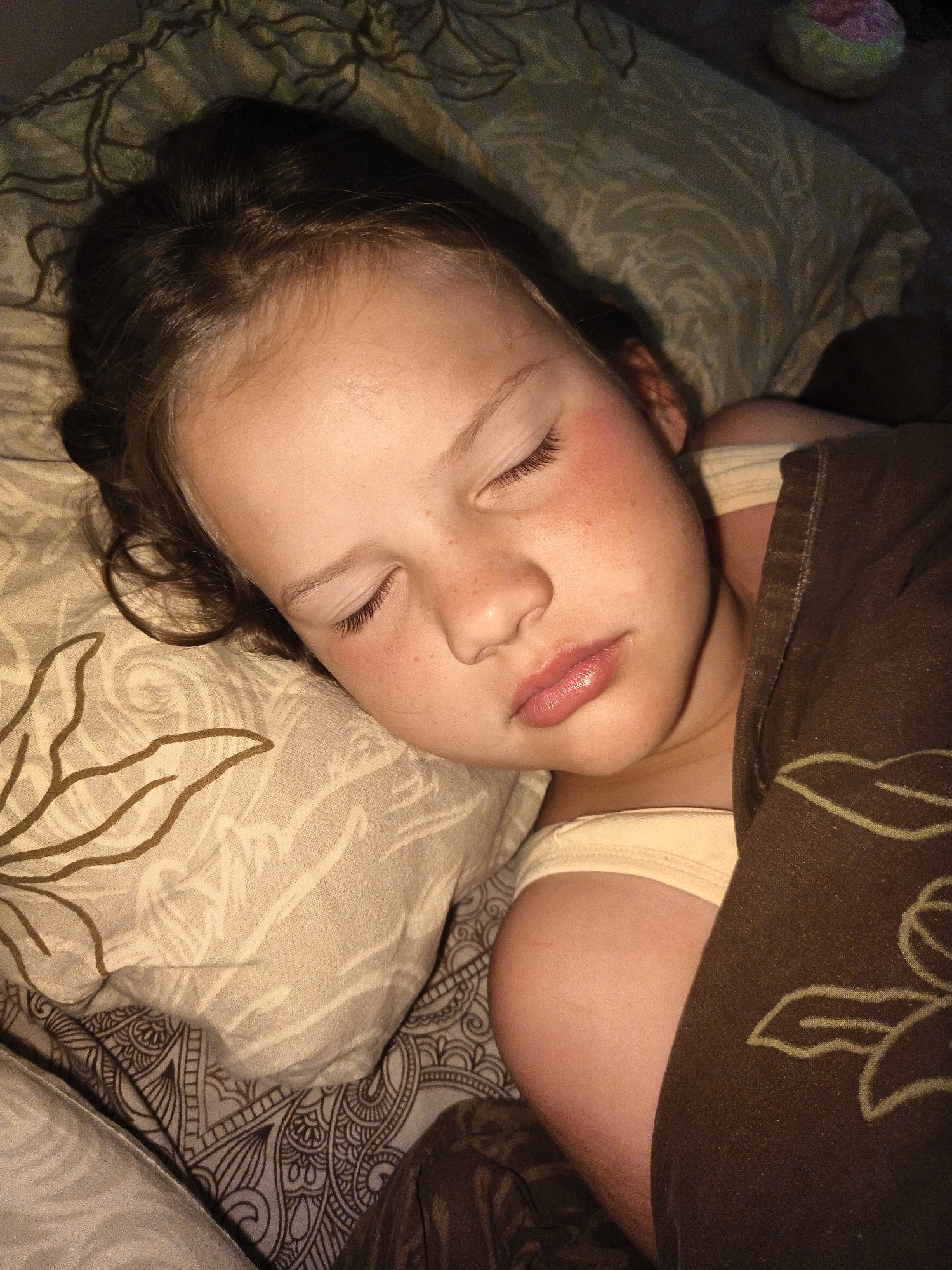 A close-up, overhead shot captures a young girl sleeping peacefully on her side. Her eyes are closed, and her face is turned slightly towards the viewer. She has fair skin with a few freckles visible on her cheeks and nose. Her lips are slightly parted, and her mouth appears relaxed. Her eyelashes are long and dark. Her hair is dark brown and falls loosely around her face and shoulders. She is wearing a light-colored tank top. A brown blanket with a floral pattern is pulled up around her, partially covering her body. She is lying on a pillow with a similar floral pattern. The background appears to be a bed with patterned sheets. The lighting is dim, suggesting it is either nighttime or the room is dark. There are no visible text elements in the image.