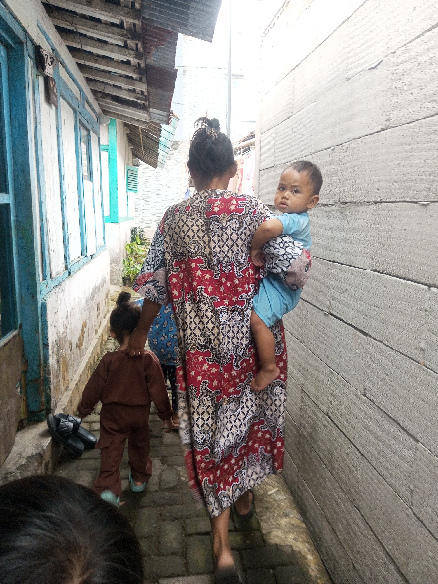 In a narrow outdoor alleyway in Andongsili, Indonesia, a woman in a long, patterned batik dress carries a toddler on her back. The woman's hair is tied up with a clip. The toddler, wearing a blue shirt and shorts, looks towards the camera. To the woman's left, a smaller child in a brown outfit walks ahead, facing away from the camera. A pair of black sandals rests on a ledge near the smaller child. The alley walls are constructed of rough, weathered wood on the right and painted wood with blue window frames on the left. The ground is paved with stone bricks. A tin roof is visible at the top of the alley.