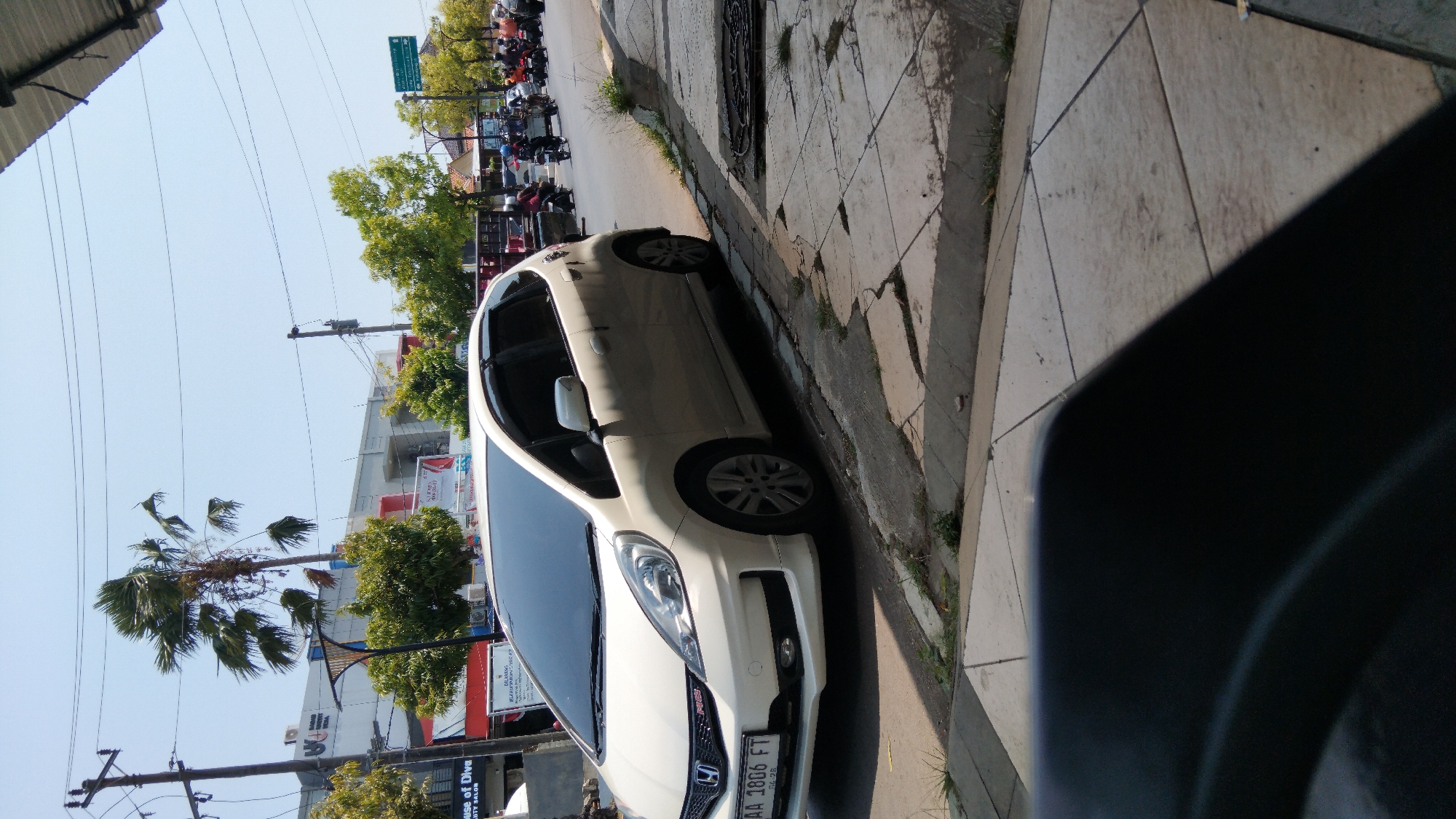 The image shows a white Honda Jazz parked on the side of a street in Batang, Indonesia. The car is parked on a concrete sidewalk adjacent to a paved road. In the background, there are buildings, trees, and utility poles. A group of motorcycles and scooters are seen on the road, indicating traffic. The sky is clear and blue, suggesting a sunny day. The license plate of the car reads "AA 1806 FT". The foreground is partially obscured by a dark object, possibly the seat of a vehicle from which the photo was taken.