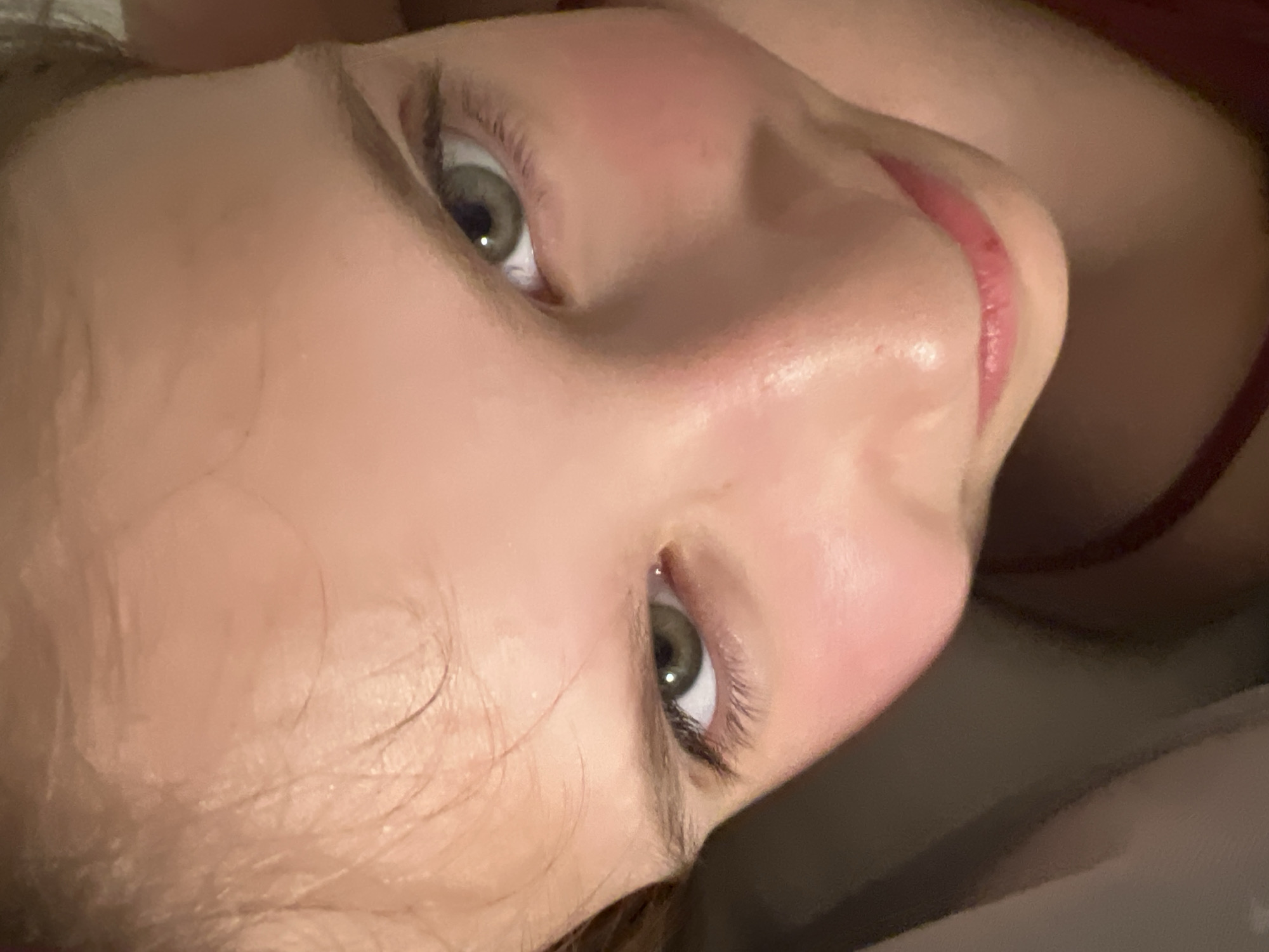 A close-up, angled portrait features the face of a young person, likely female, from the forehead to the chin. The subject appears to be lying down, looking directly upwards into the camera with a neutral, relaxed expression. The skin is fair with subtle pink undertones visible on the cheeks and around the nose. Both eyes are open and clearly visible, showcasing light green or hazel irises, distinct pupils, and long, dark eyelashes. A straight nose is prominent in the center of the face. The lips are a soft pink, slightly parted, and show natural texture. Wispy strands of light brown hair are visible around the forehead and temples. On the right side of the image, a thin red strap, suggestive of a top or camisole, is partially seen. The background is dark, soft, and out of focus, indicating an indoor setting. The lighting is gentle, illuminating the facial features. No activity is explicitly depicted beyond posing for the photograph. There is no visible text. The scene conveys a calm and direct gaze.
