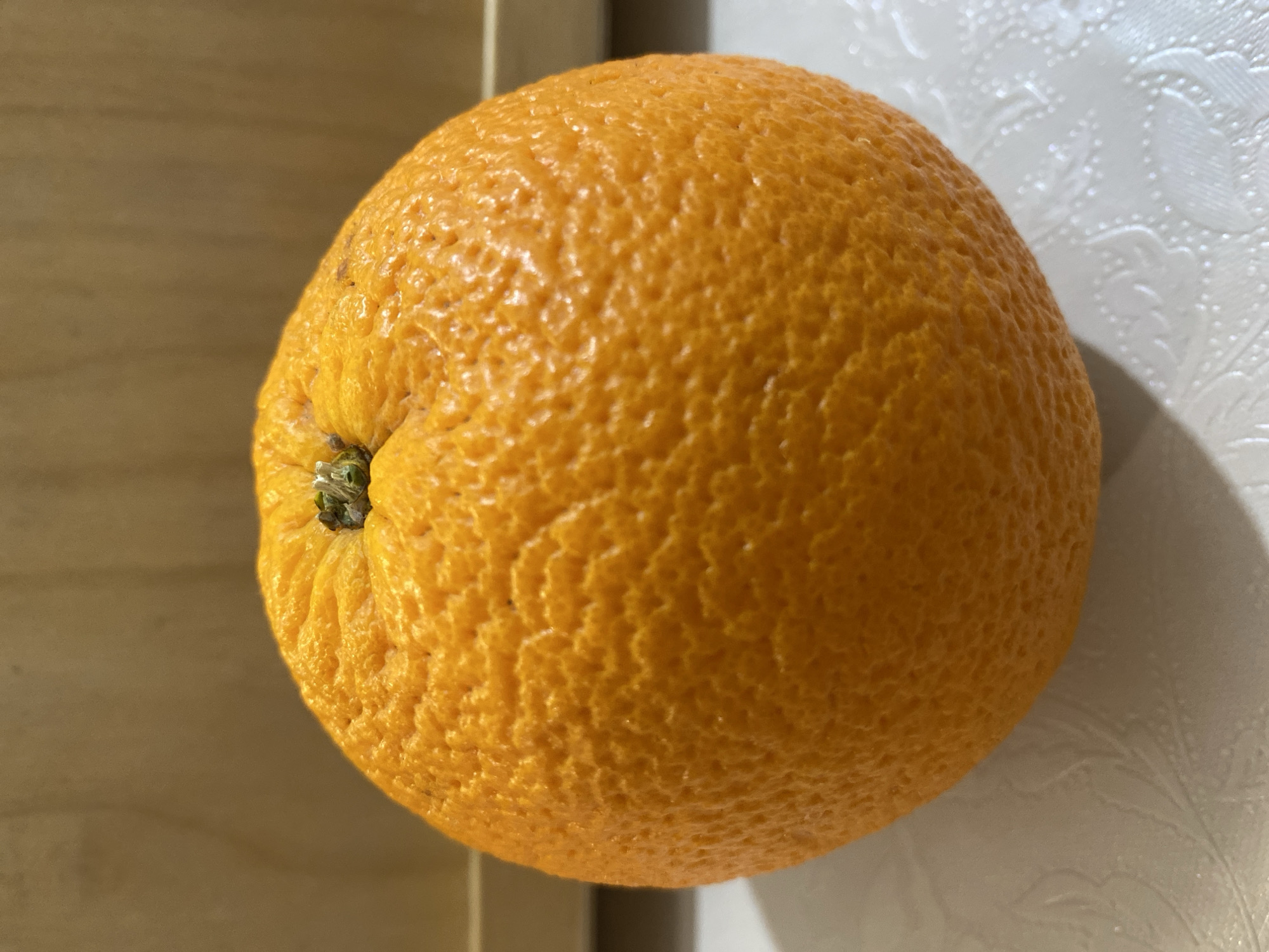 A top-down, close-up shot depicts a whole orange resting on a surface. The orange is round with a textured, orange peel. The stem area, located on the left side of the image, displays some green remnants. The surface beneath the orange is split between a beige, wooden-like material on the left and a white surface with subtle patterned indentations on the right. The scene is brightly lit.
There is no information about Budva, Montenegro, in the image.