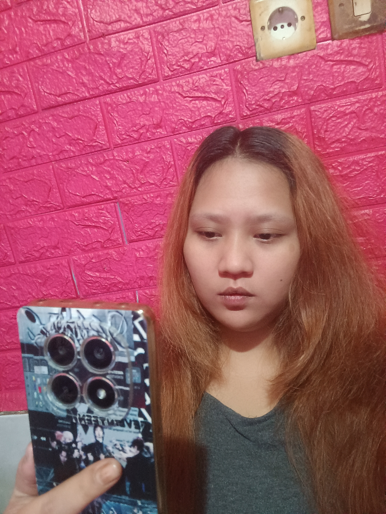 A young woman with light brown hair is taking a selfie against a bright pink brick-patterned wall. She is wearing a grey t-shirt and is holding a smartphone with a decorative case featuring what appears to be a group of people. There are two electrical outlets visible on the wall behind her. The image appears to be taken indoors in a room.