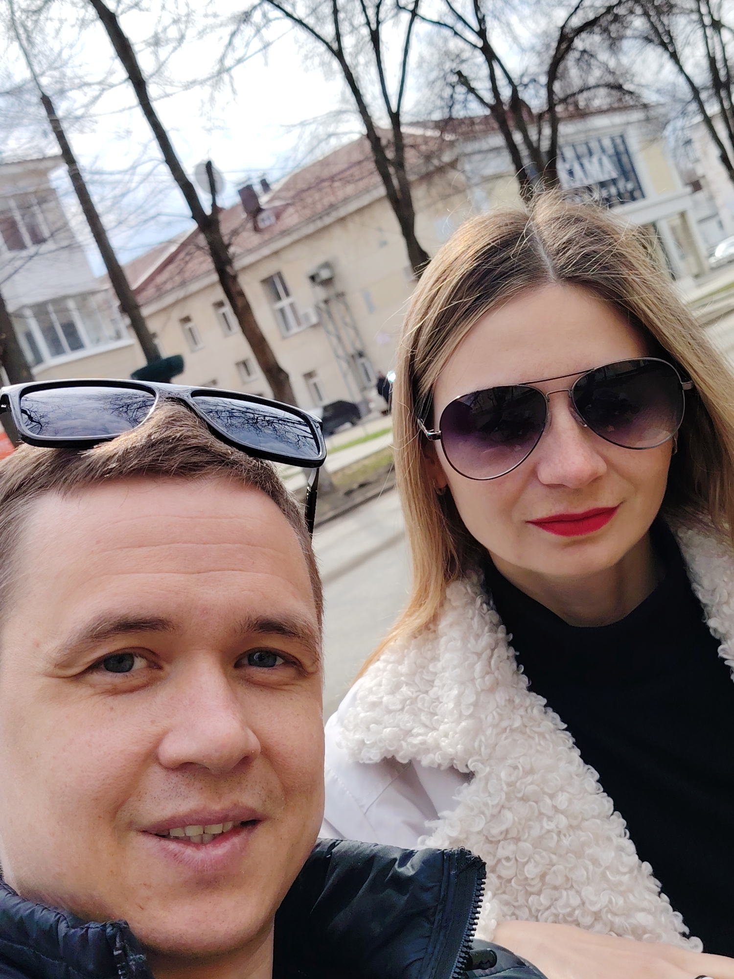 The image depicts a selfie of two individuals, a man and a woman, likely taken outdoors in a city, evidenced by the presence of buildings and trees in the background. The man is positioned on the left side of the frame, looking directly at the camera with a slight smile. He has short, light brown hair with sunglasses placed on top of his head. He is wearing a dark jacket. The woman is on the right side of the frame, also wearing sunglasses and red lipstick. She is wearing a black top and a white, textured coat. The background shows buildings and bare trees, indicating a potentially urban setting in the city of Chaikovsky, Russia. The lighting suggests it is daytime.