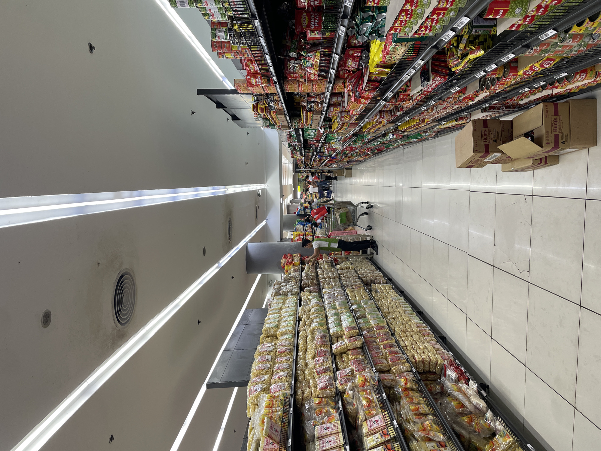 The image shows an aisle in a supermarket, likely in Davao City, Philippines, with shelves stocked with various food products. On the left, rows of packaged noodles are displayed, and on the right, shelves are filled with canned goods, including what appear to be vegetables and other food items. Several shoppers and supermarket employees are present in the aisle. One employee, wearing a green vest, is stocking shelves on the left side. A shopping cart is visible near the center of the aisle. The lighting is provided by long, fluorescent ceiling lights. The floor is tiled and appears to be clean. Boxes of products are stacked on the floor near the shelves on the right side. The overall scene depicts a typical retail environment within a supermarket.