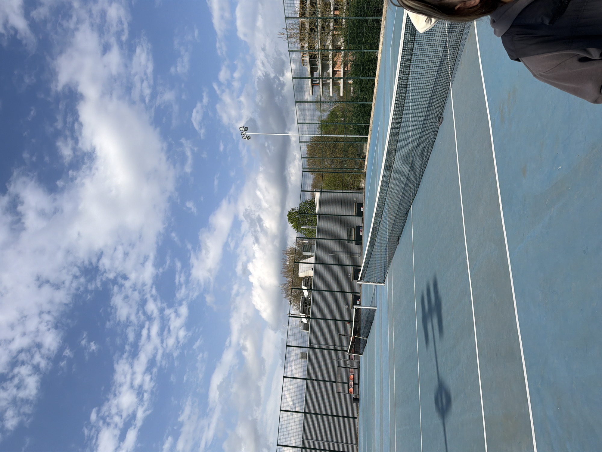 The image shows a blue tennis court with white lines, a net running across the middle. The court is enclosed by a chain-link fence. Beyond the fence are buildings and trees. In the sky, there are scattered clouds. The shadow of a lamppost falls across the tennis court. Part of a person’s head and shoulder are visible in the upper right corner of the image. Based on the presence of tennis courts, and typical urban planning for sport, the location is consistent with the city of Blagoevgrad, Bulgaria, but this determination cannot be confirmed.