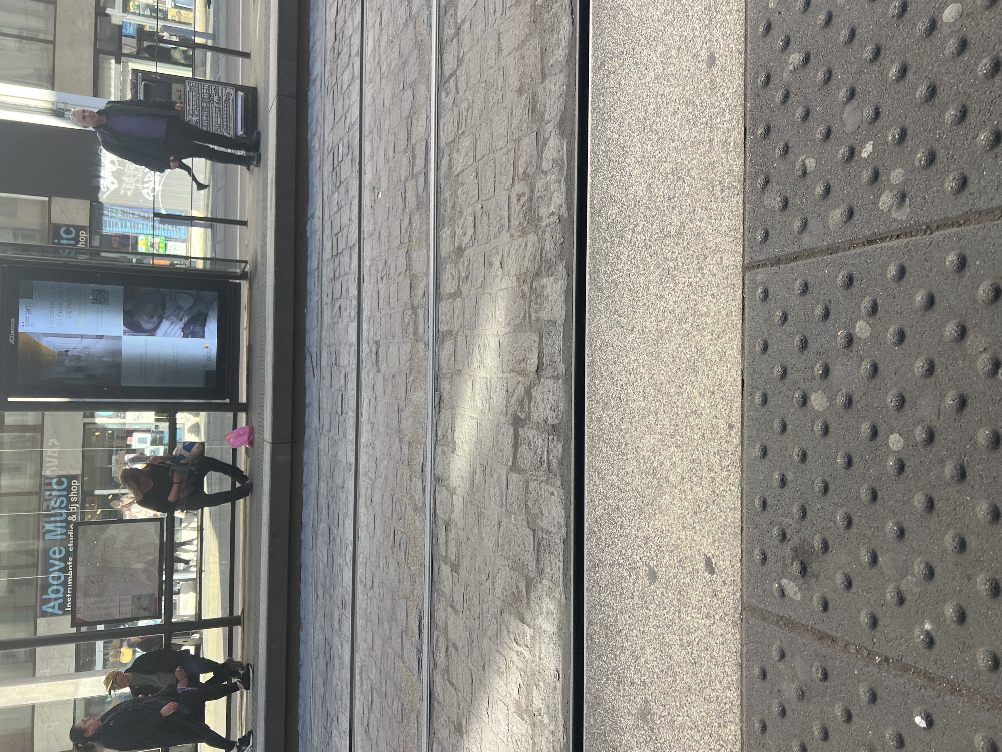 The image shows a street scene in Strasbourg, France. The foreground features textured tactile paving, a grey paved area and tram tracks. A tram stop is visible to the left with advertisements and passersby. A sign for "Above Music" is visible on the tram stop structure. People are standing and walking, waiting at the tram stop.