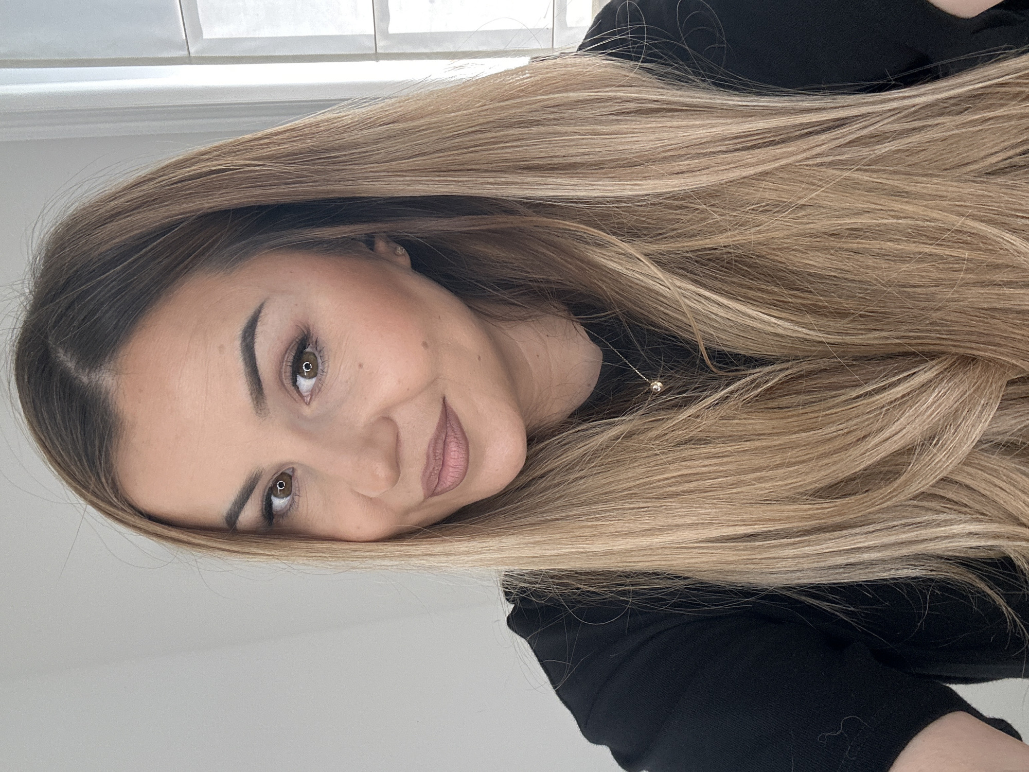 The image presents a headshot-style portrait of a single female subject, estimated to be in her late 20s to early 40s, captured indoors in Asenovgrad, Bulgaria. The subject is positioned centrally, looking directly at the viewer with a slight, closed-lip smile.
Her hair is long, extending past her shoulders, and features a multi-tonal light brown base with numerous blonde highlights, creating a natural-looking balayage or ombré effect. It is parted on the right side and appears smooth with slight waves at the ends. Her eyes are brown with distinct, circular light reflections, suggesting a frontal light source such as a ring light or window. Her eyebrows are dark, well-defined, and precisely shaped. She wears visible eye makeup, including eyeliner and mascara, and her lips are colored with a matte, muted pink or nude lipstick. A small beauty mark is located below her right eye.
The subject is dressed in a plain black, long-sleeved top, likely made of a fine knit or jersey fabric. She wears a delicate silver-toned necklace with a small, circular, light-colored pendant, and a single small stud earring is visible in her left ear.
The background is simple and uncluttered, consisting of a plain, light gray or off-white wall on the left side of the frame. To the right, a vertical white element, consistent with a window frame or door jamb, is visible, indicating an indoor setting with ambient light.