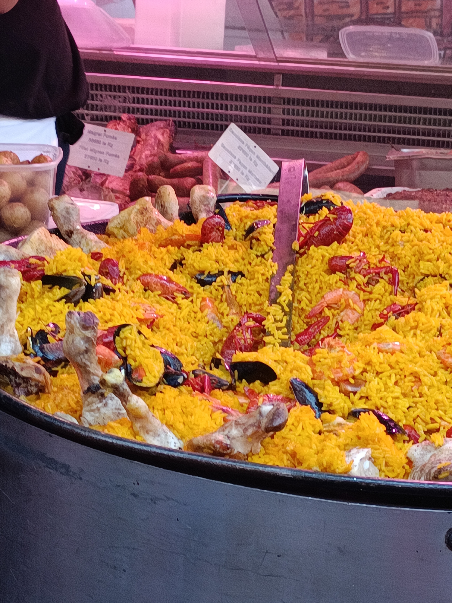 A large pan of paella is displayed at a market stall in Le Neubourg, France. The paella is a vibrant yellow color and is filled with rice, shrimp, crayfish, mussels, and pieces of chicken or rabbit. The pan is deep and wide, and the food is piled high. In the background, there are refrigerated display cases containing various meats, including sausages and cuts of beef or pork. A sign is visible near the meat display, which appears to be a price list. A person wearing a black sweater and a white apron is partially visible on the left side of the image, attending to the stall. The lighting in the market is bright, with pinkish tones reflecting off the displays and ceiling.