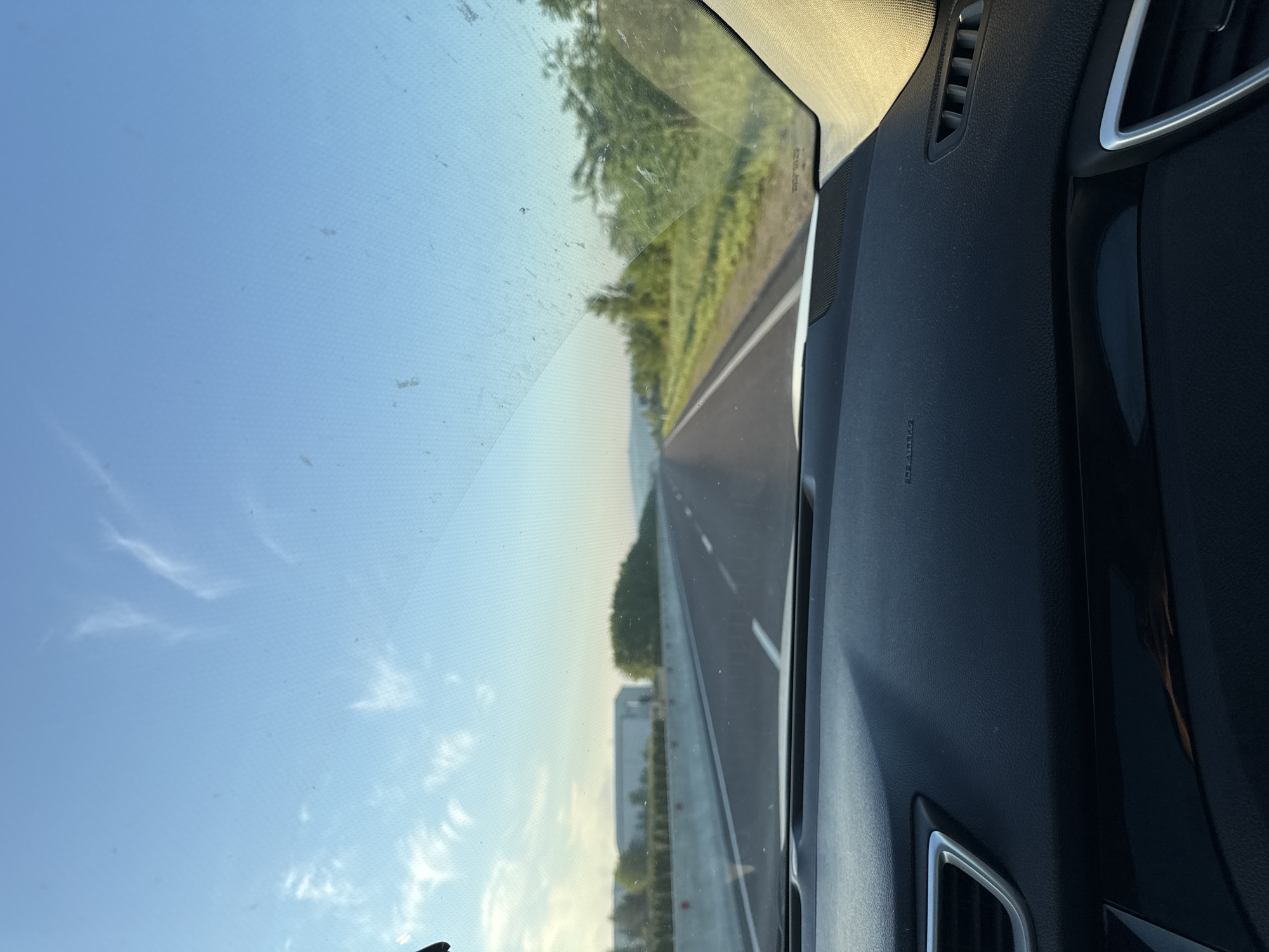The image is a first-person view from inside a car driving on a highway in Cojușna, Moldova. The road stretches into the distance, flanked by green fields and trees on either side. A concrete barrier runs along the left side of the highway, with a large building and more trees visible beyond it. The sky is clear blue with a few wispy clouds. The car's dashboard is visible in the foreground, with the words "SRS AIRBAG" printed on it.